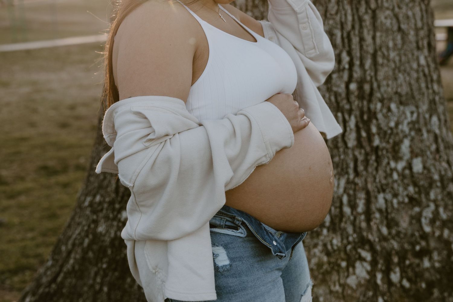 Side view of pregnant person wearing white sweater and jeans leaning against tree.