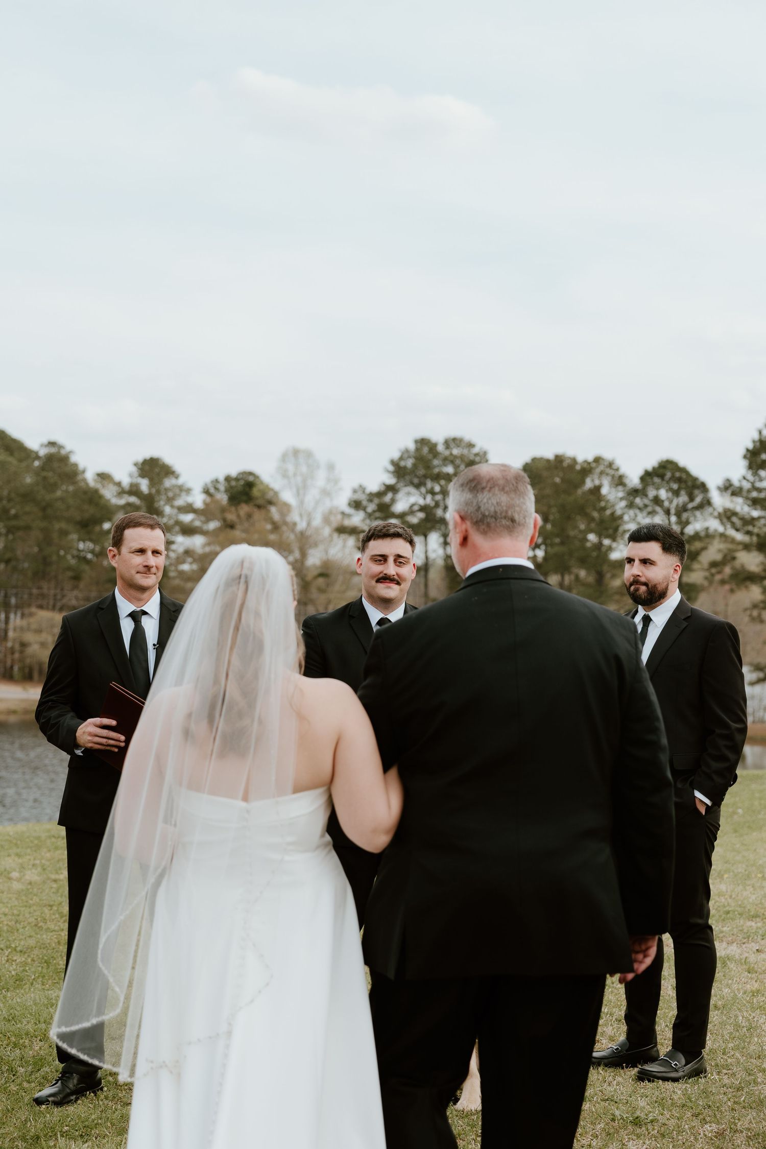 Wedding party members in black suits gather outdoors on a grassy field.