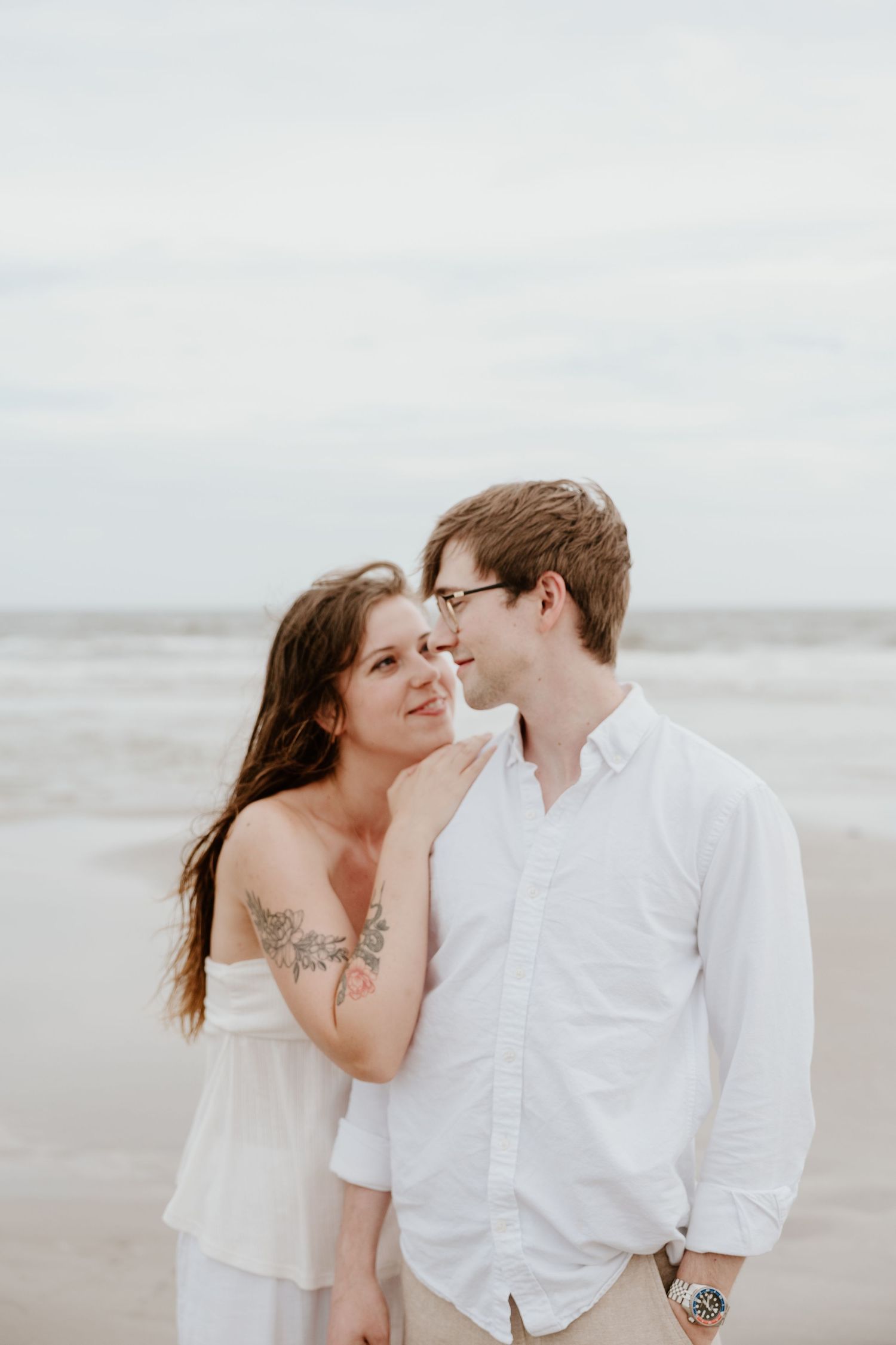 Casual beach photoshoot of couple in white outfits sharing intimate moments by the ocean.
