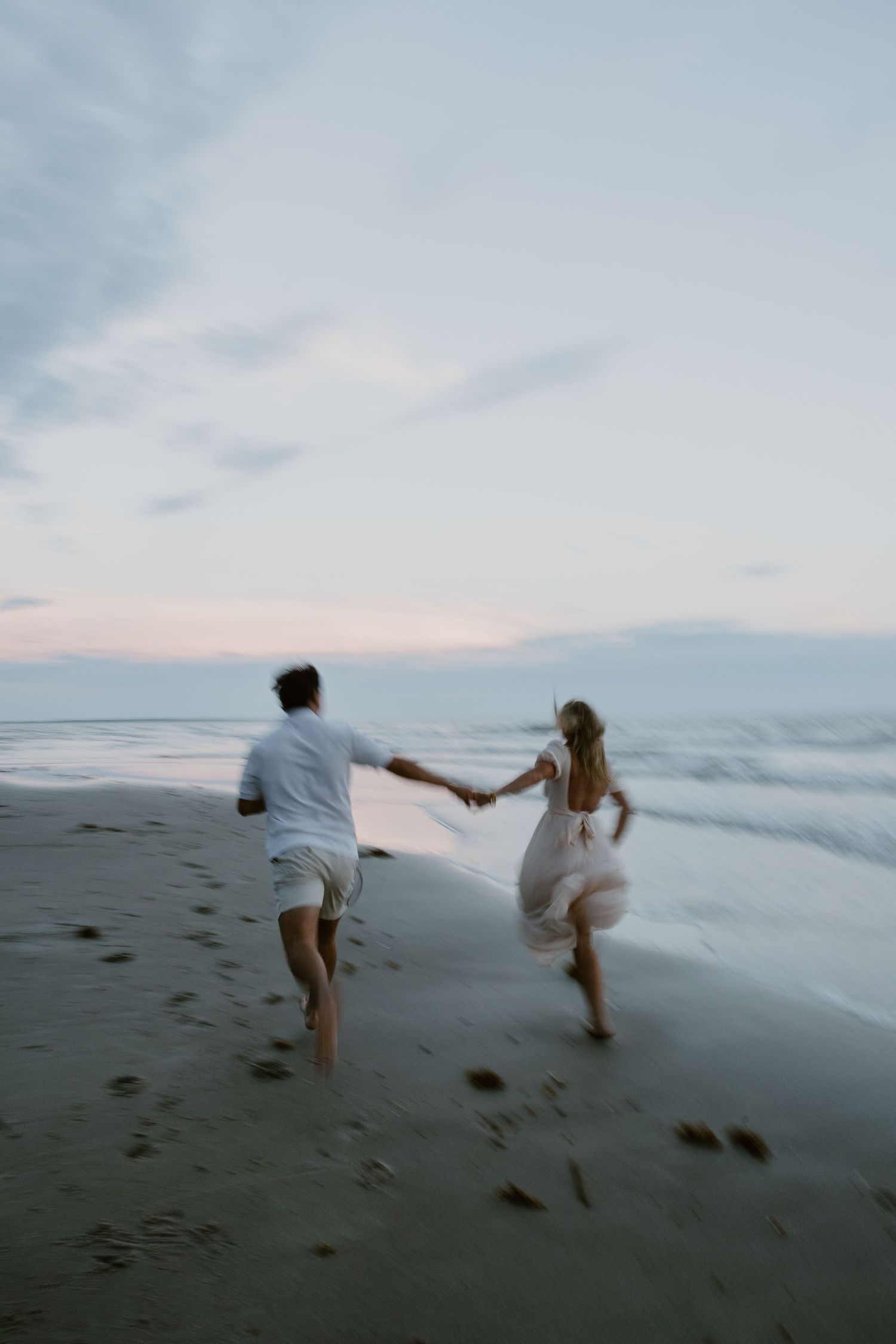Couple running playfully along beach shoreline at dusk holding hands.