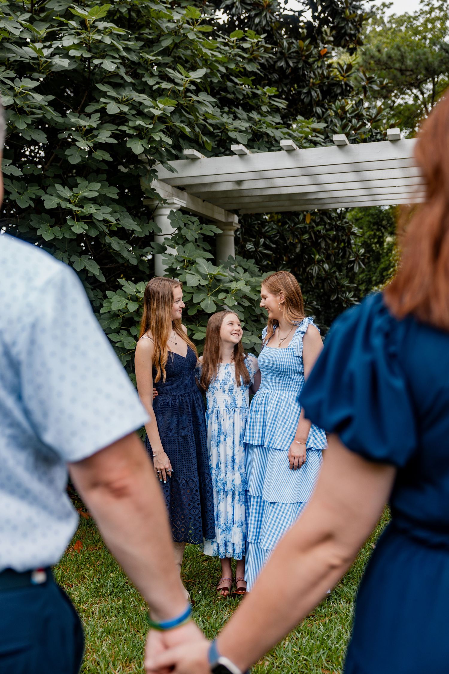 Group gathered in garden setting wearing coordinated blue dresses near white pergola.