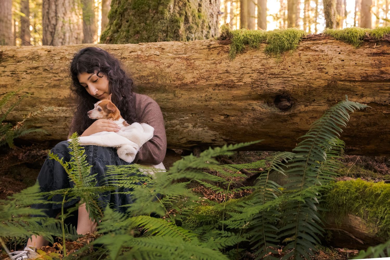 Woman sitting against a fallen log in a forest, tenderly holding an orange and white cat among green ferns.