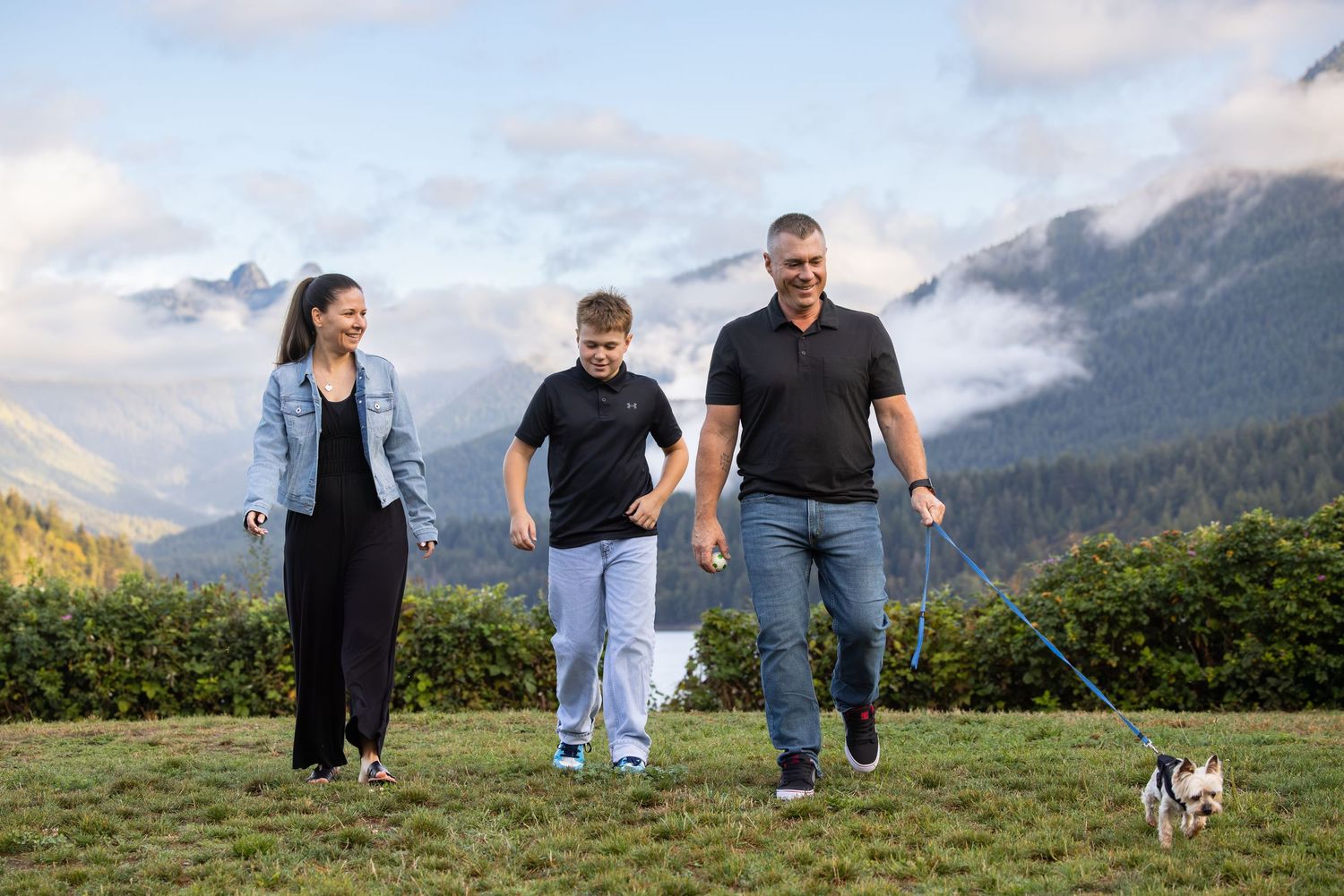 Family of three walking a small dog on a leash in a green mountain landscape with cloudy skies.