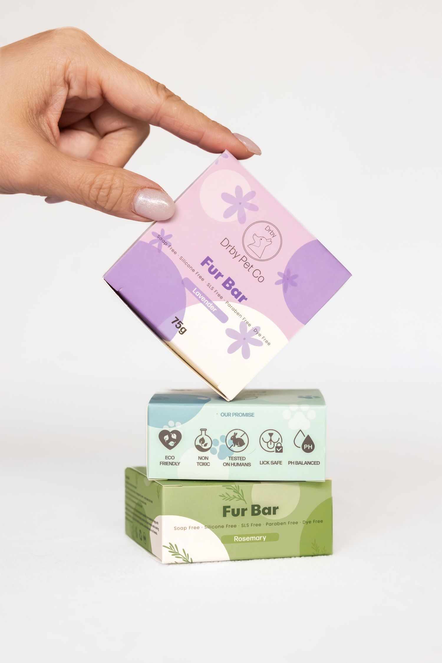 Hand holding Dry Pet Co Fur Bar soap box above two stacked Fur Bar pet shampoo bars in purple and green packaging.
