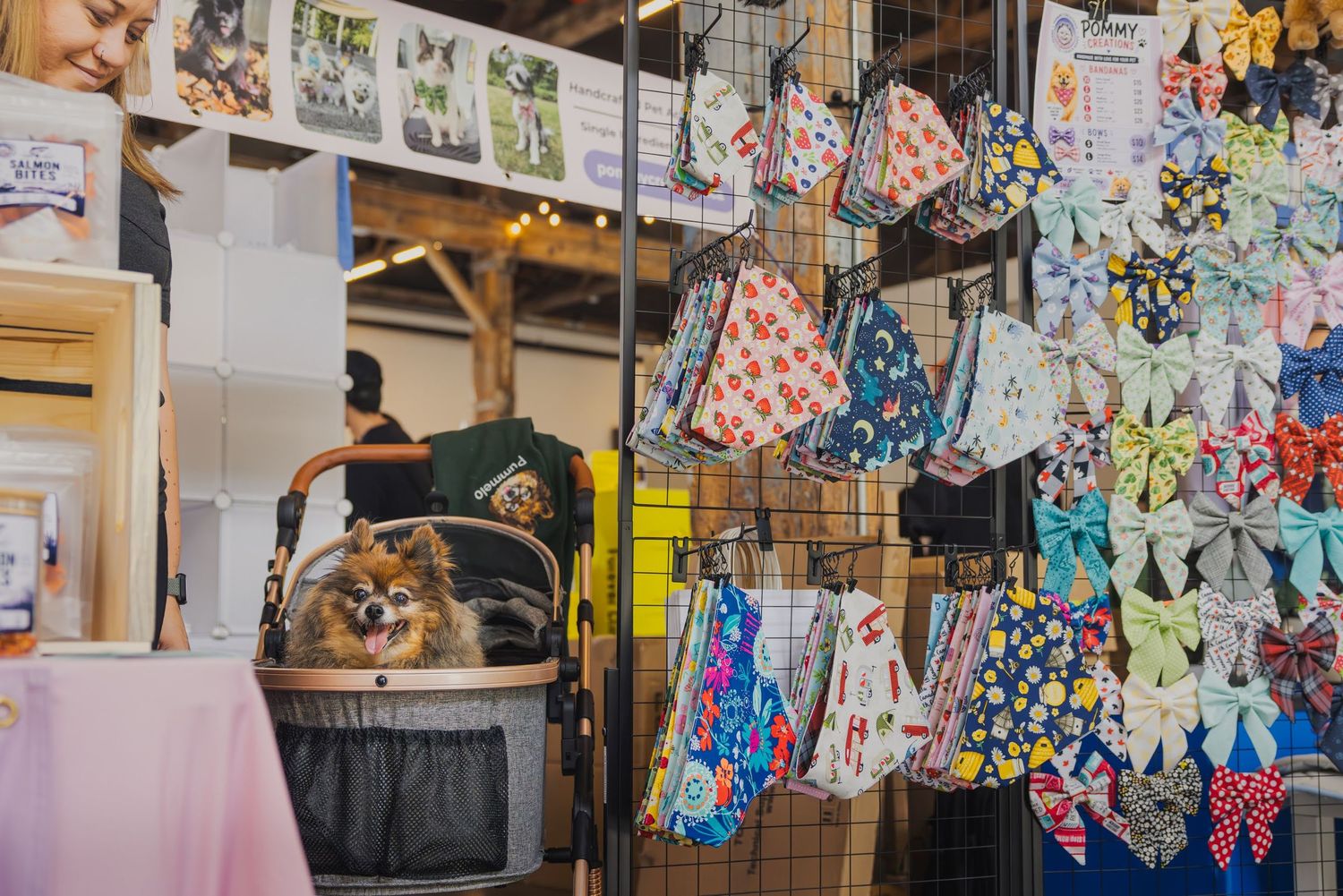 A fluffy Pomeranian in a basket beside colorful pet bandanas and bow ties displayed at a pet accessories market booth.