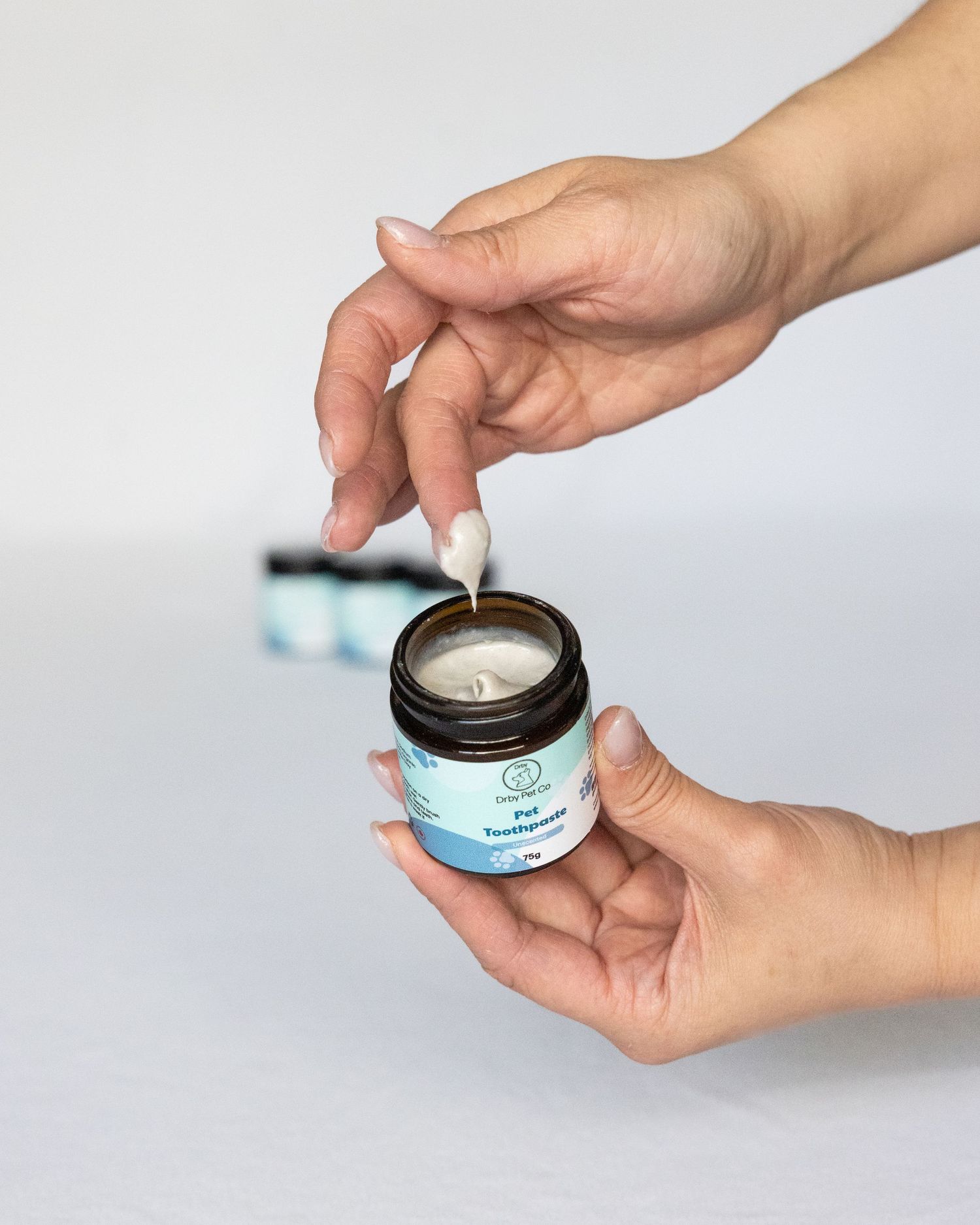 Hands holding an amber jar of Pet Toothpaste with a small applicator being dipped into the product.