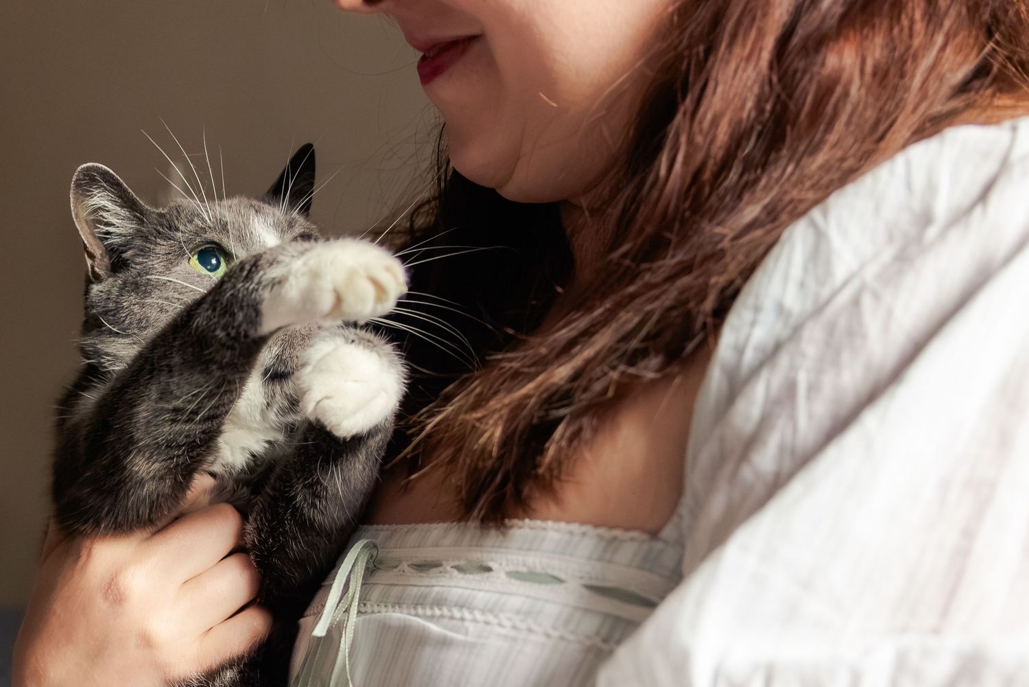 A woman in a white top holds a gray and white cat with bright green eyes close to her face.