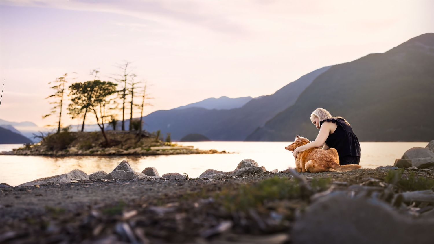 A woman sits on rocky shore petting a golden dog beside a serene mountain lake at sunset.