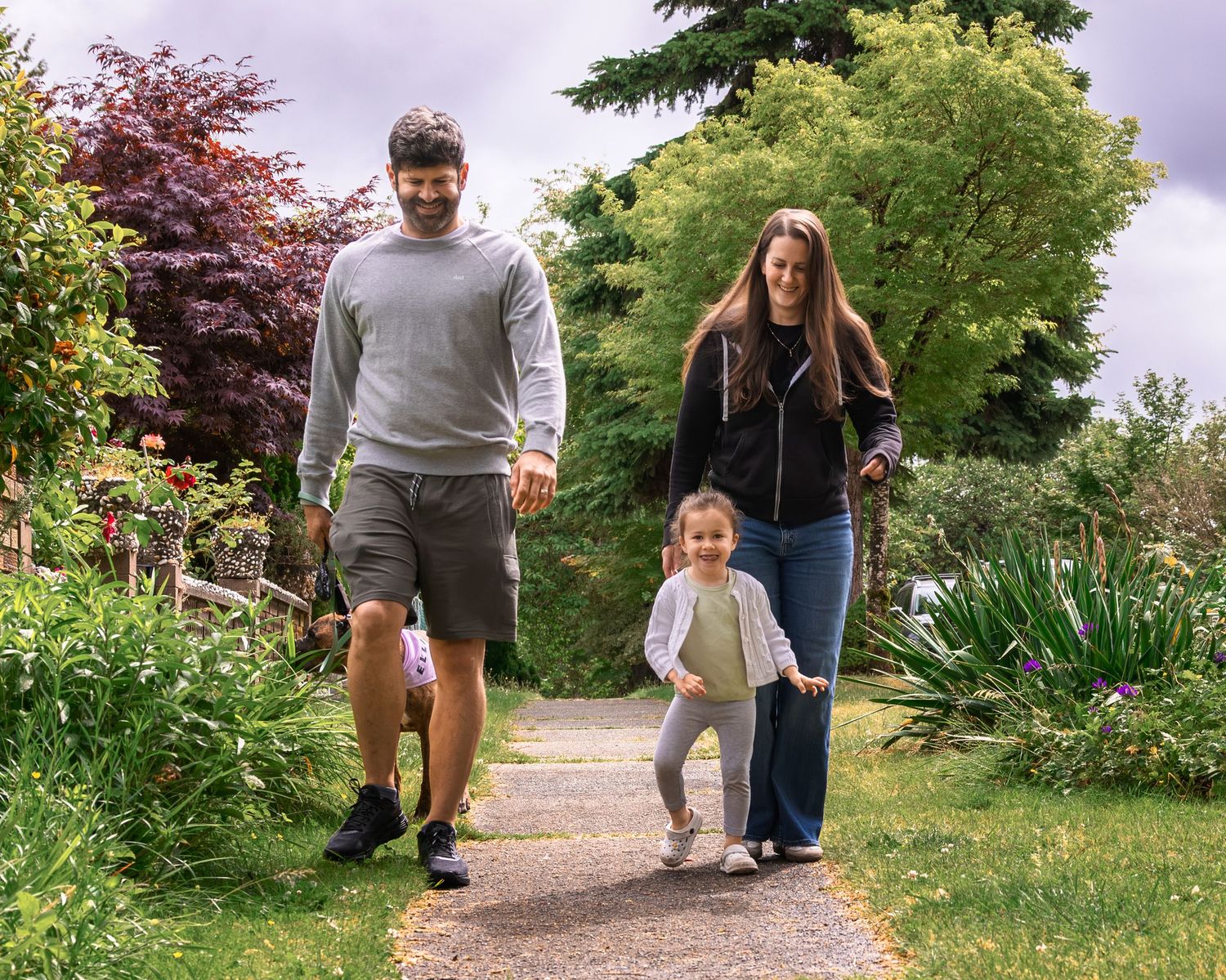 Family with young child walking together along a garden path surrounded by lush green trees.