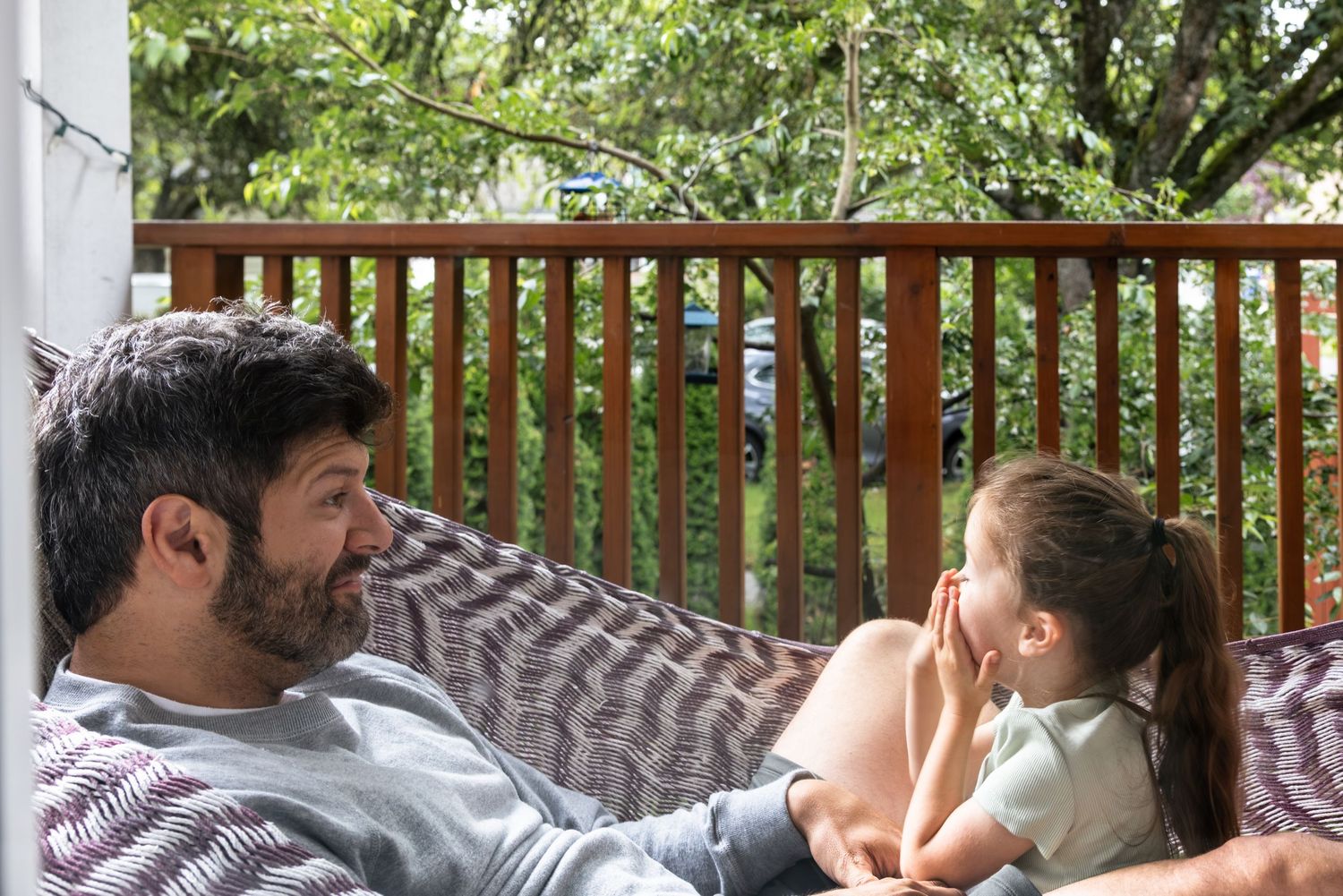 Father and daughter relaxing together on a hammock on a wooden deck surrounded by lush green trees.