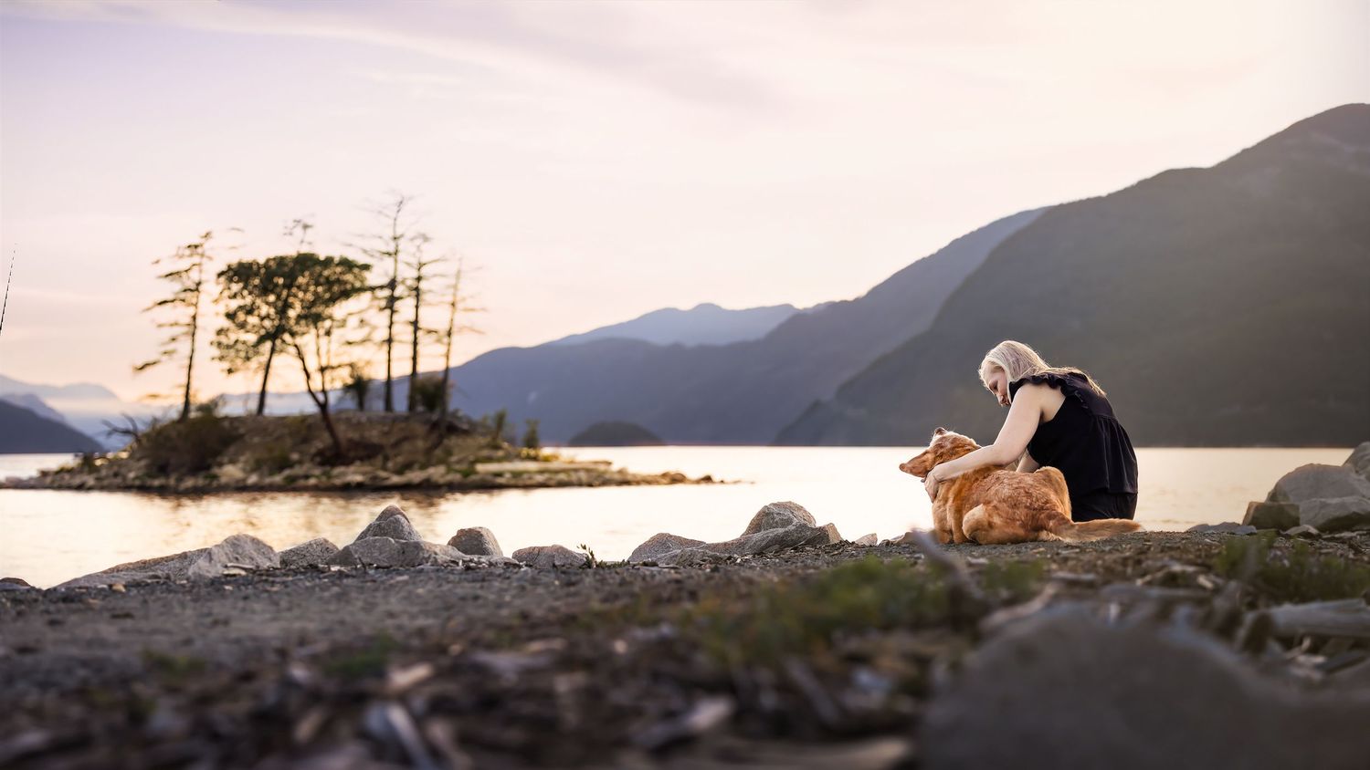 A woman sits on rocky shore petting a golden dog beside a serene mountain lake at sunset.