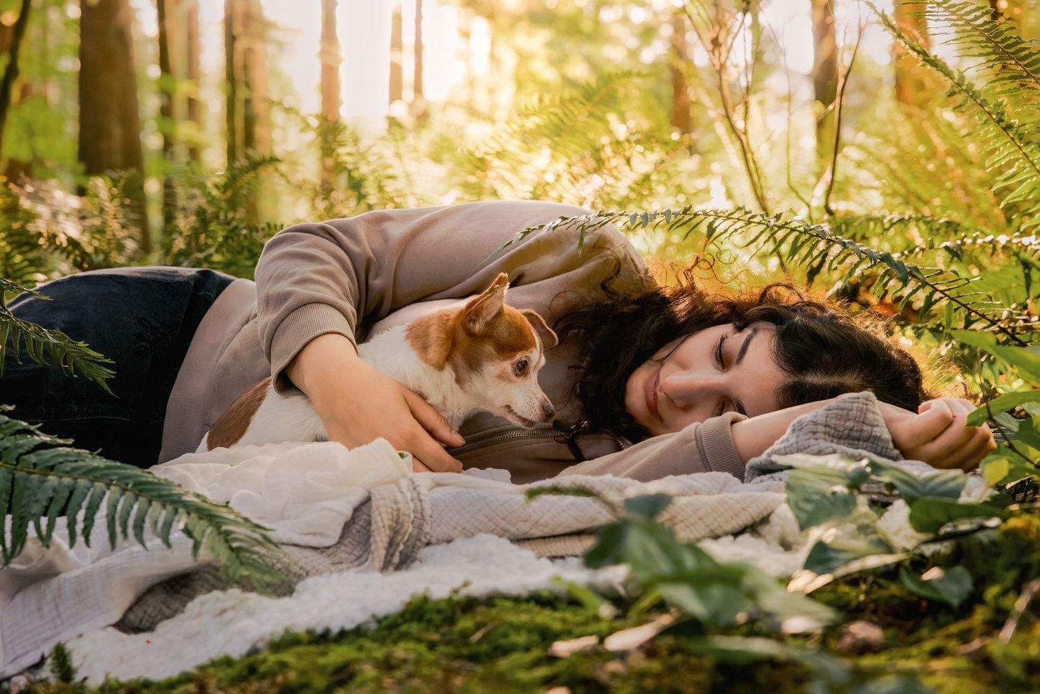 Woman lying on a blanket in a sunlit forest, cuddling with a small dog among green ferns.