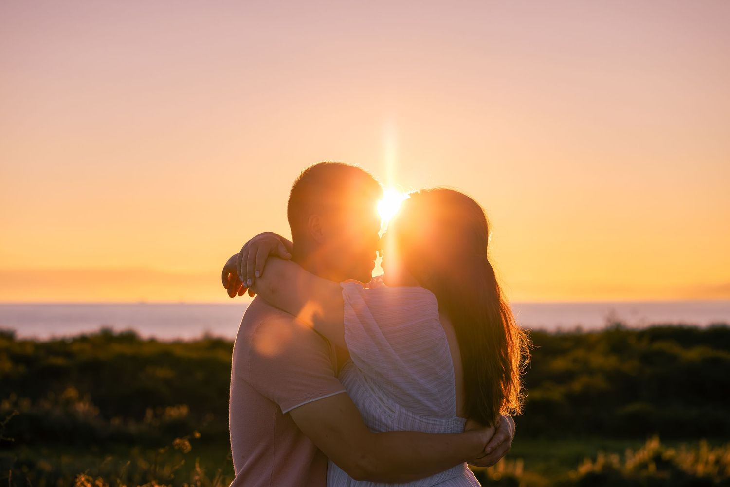 Romantic couple embracing and kissing at sunset with golden light and ocean horizon in the background.
