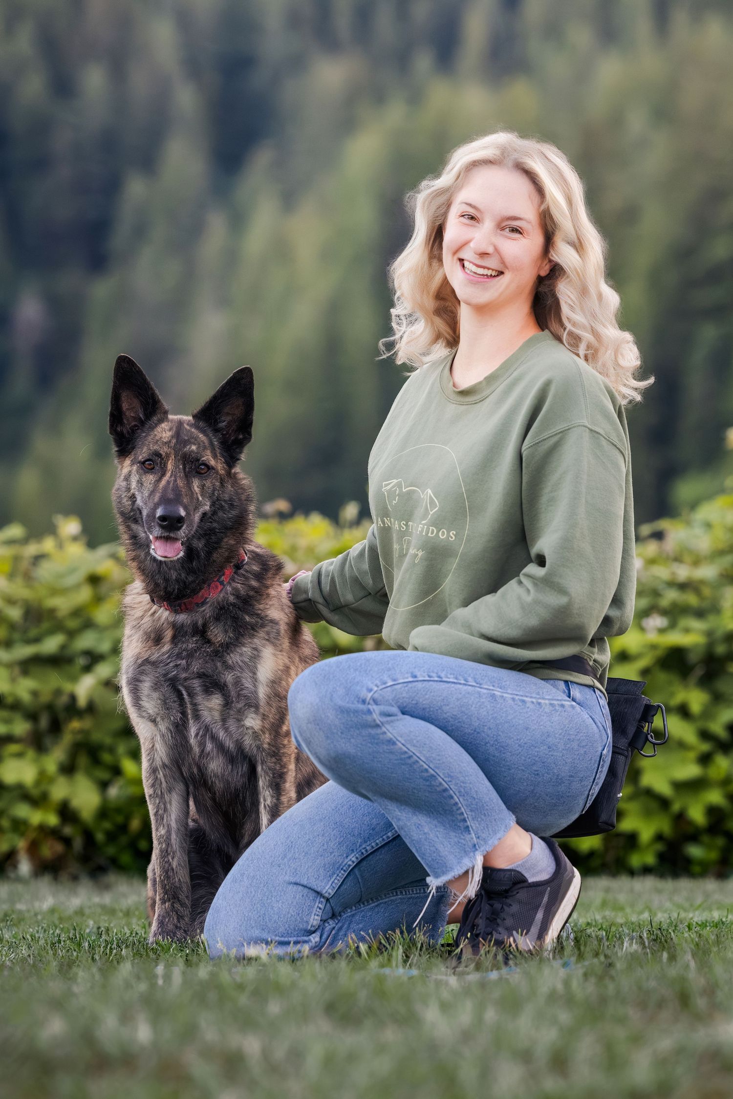 Smiling blonde woman in green sweatshirt kneeling on grass beside a German Shepherd dog with forest background.