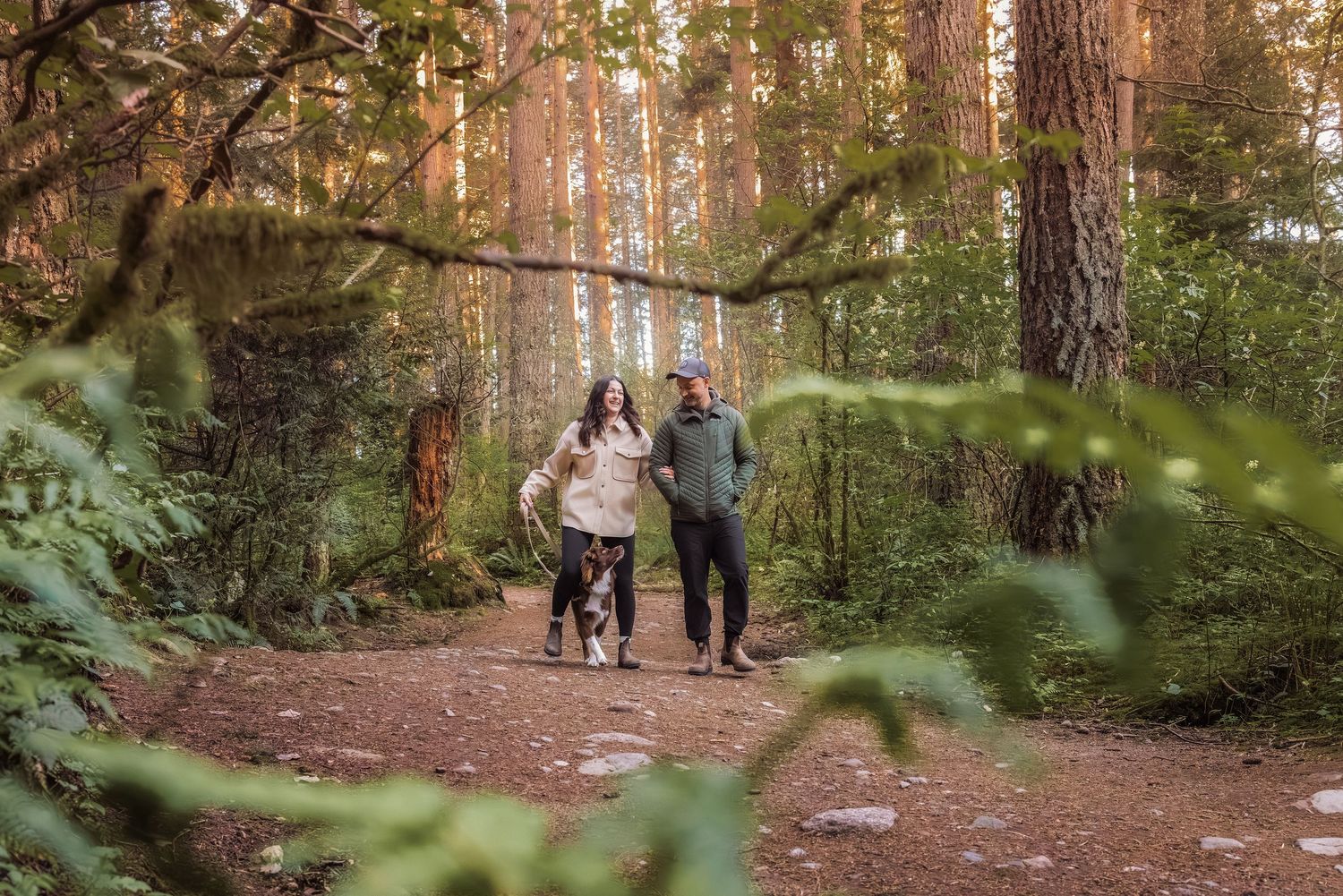 Couple walking with dog on forest trail surrounded by tall pine trees and lush green foliage.