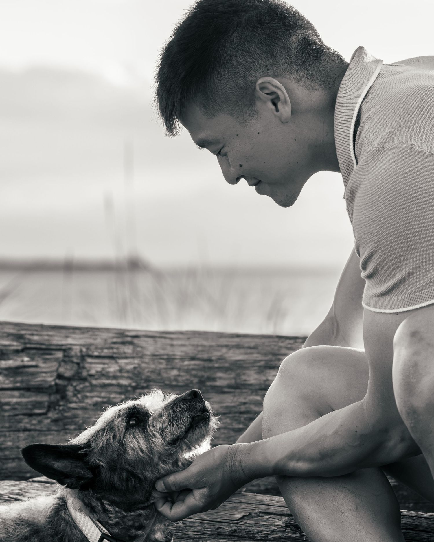 Black and white photo of a young man smiling and bonding with his dog outdoors near water.