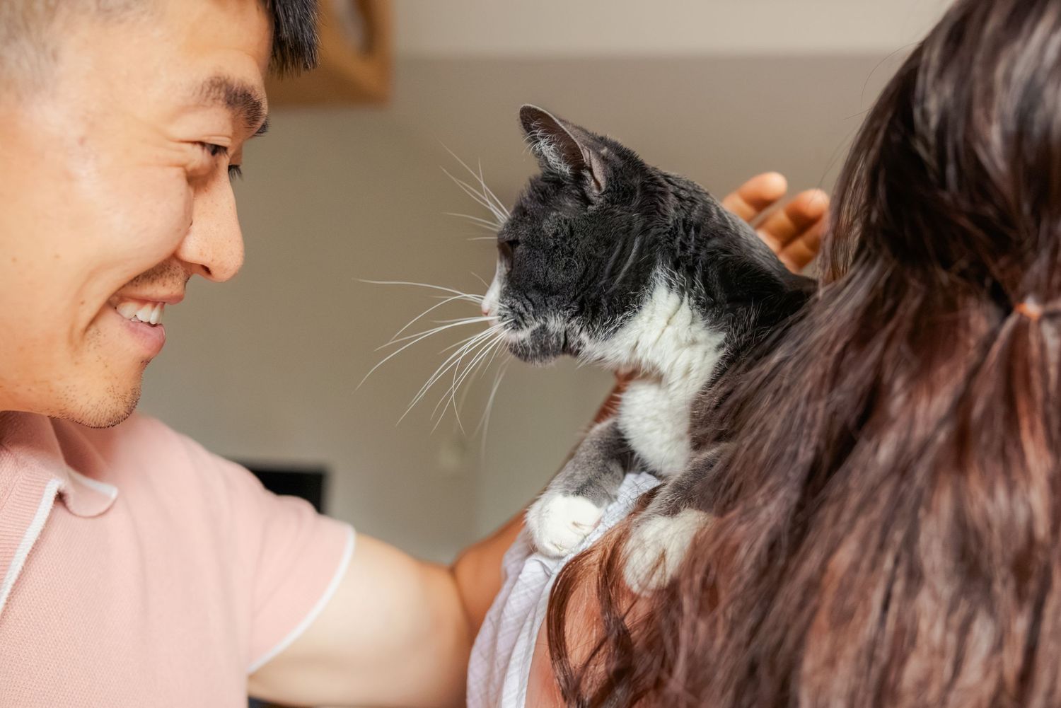 A smiling man holds a black and white cat on a woman's shoulder indoors.