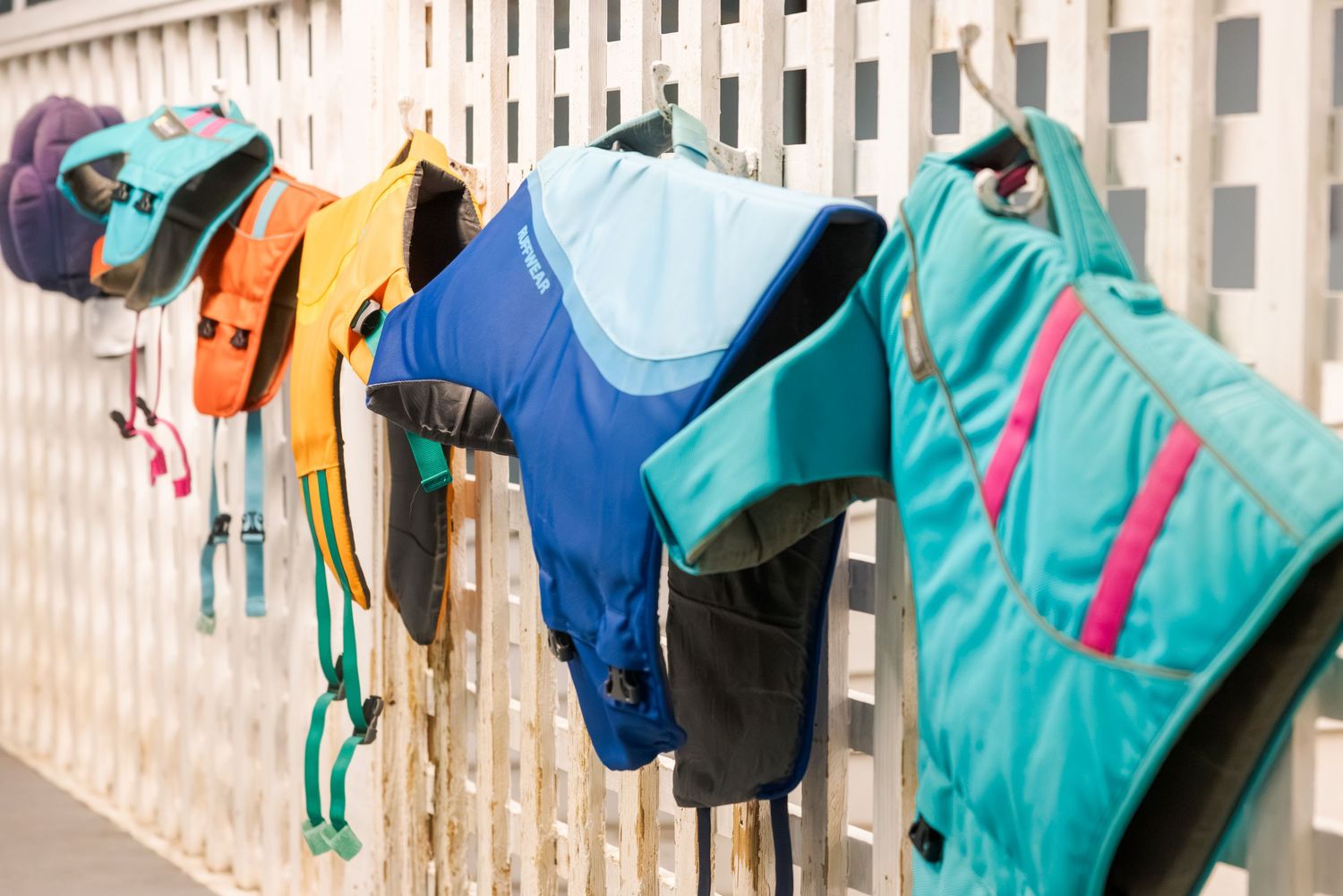 Colorful life jackets and vests hanging on a white wooden fence outdoors.