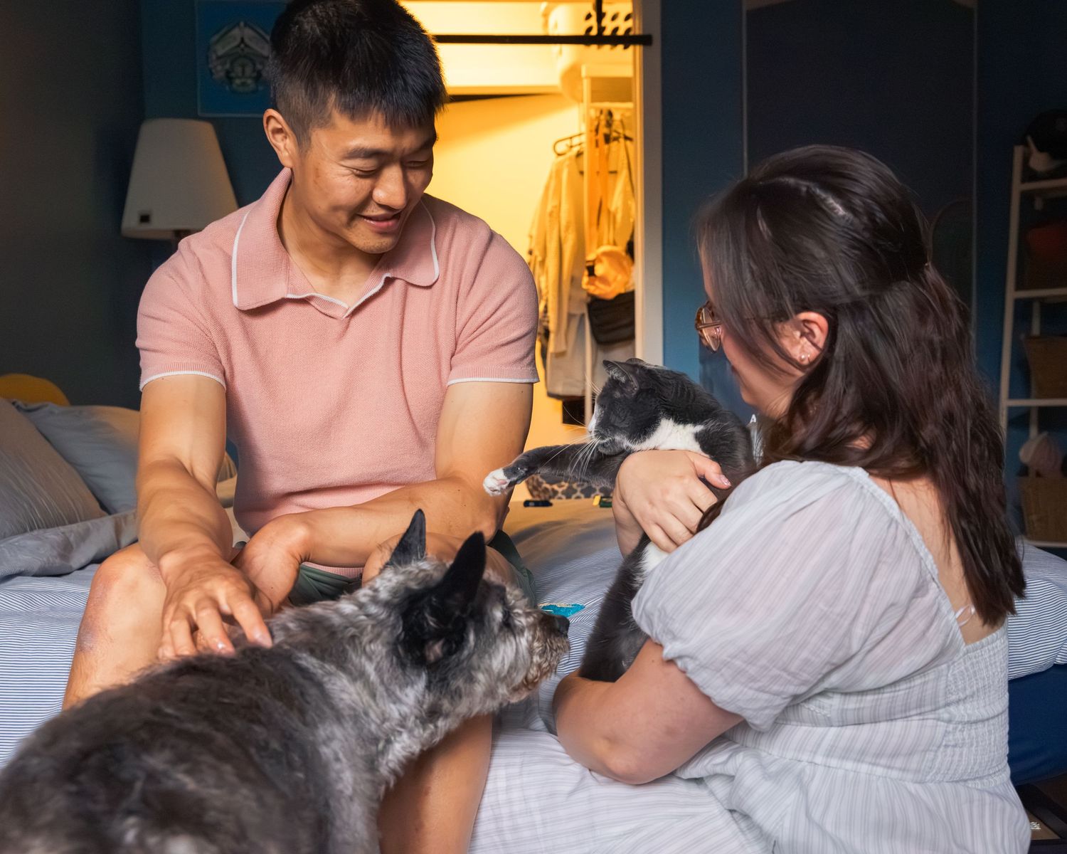 Man and woman sitting on bed playing with two cats, a gray and a black and white, in a dimly lit bedroom.