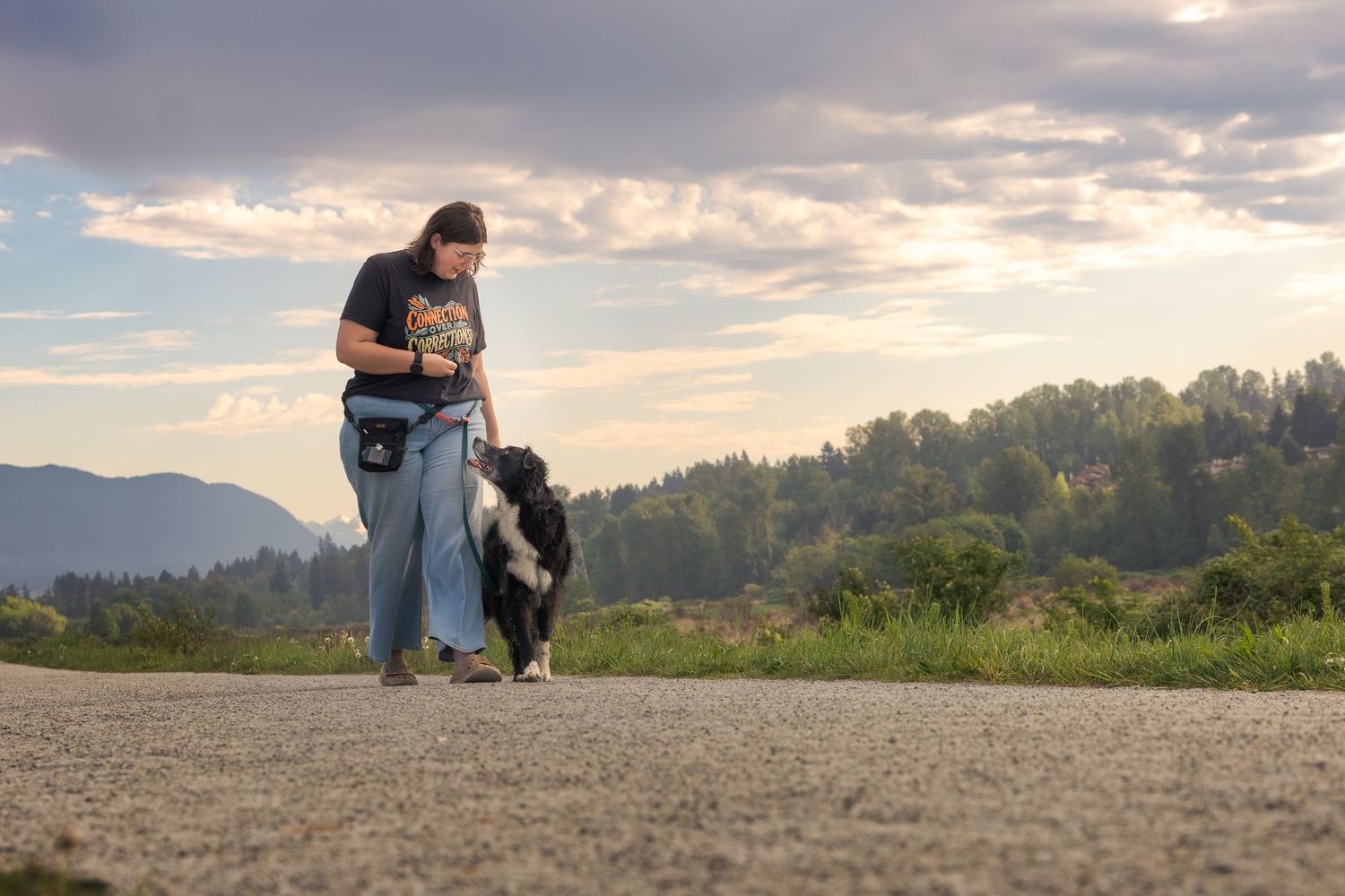 Woman training a Border Collie dog on a scenic mountain road at sunset with cloudy sky.