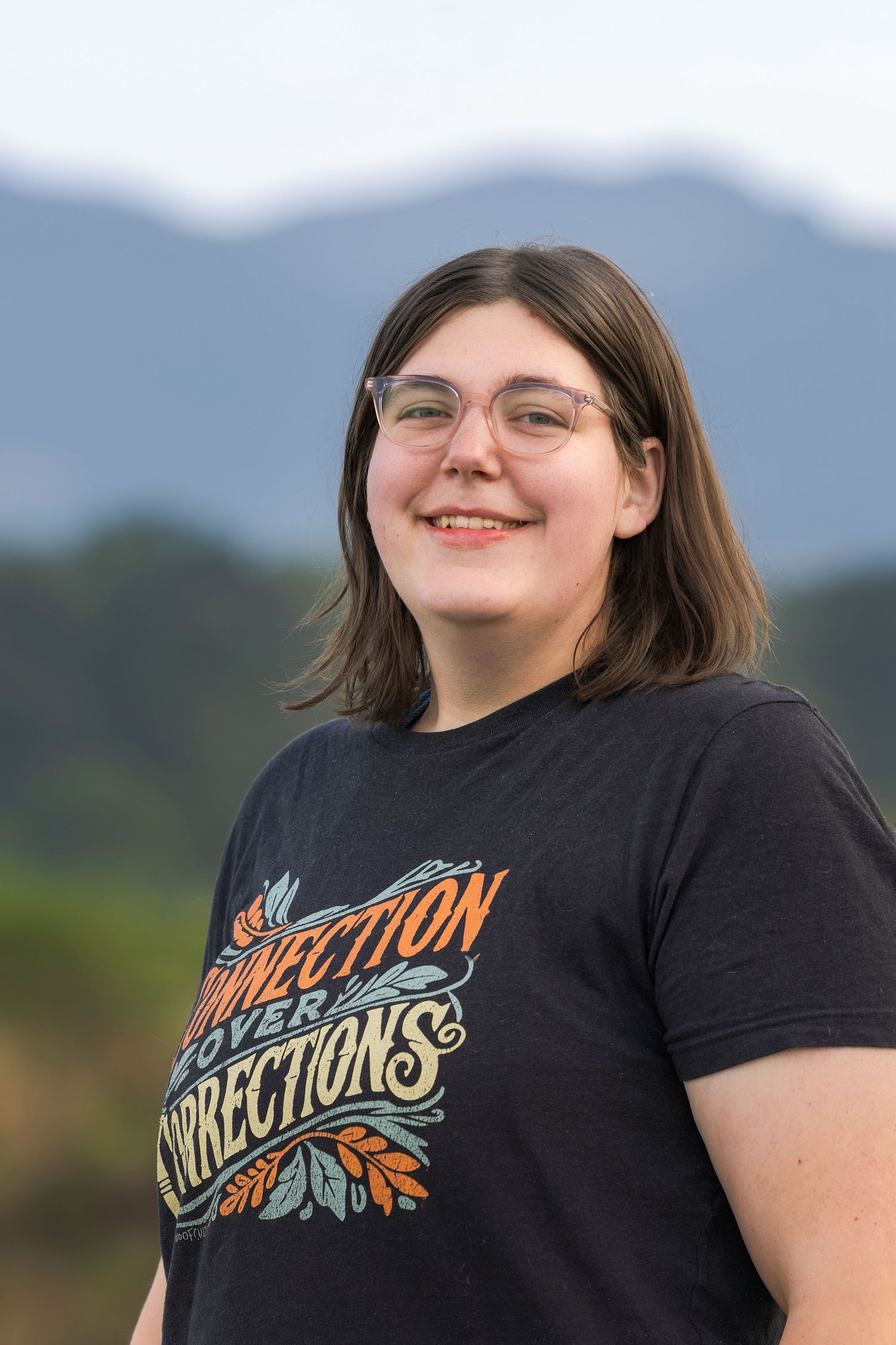 Young woman with glasses smiling outdoors, wearing a Connection Over Corrections t-shirt with mountains in background.