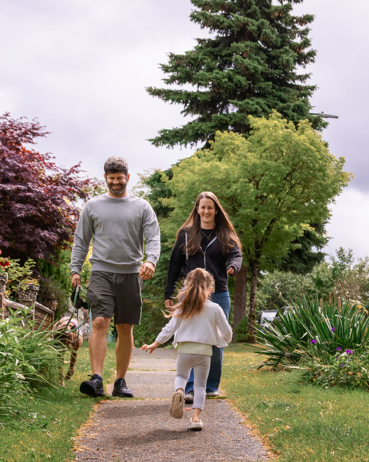 A happy family walks along a garden path surrounded by lush greenery and tall trees on a cloudy day.