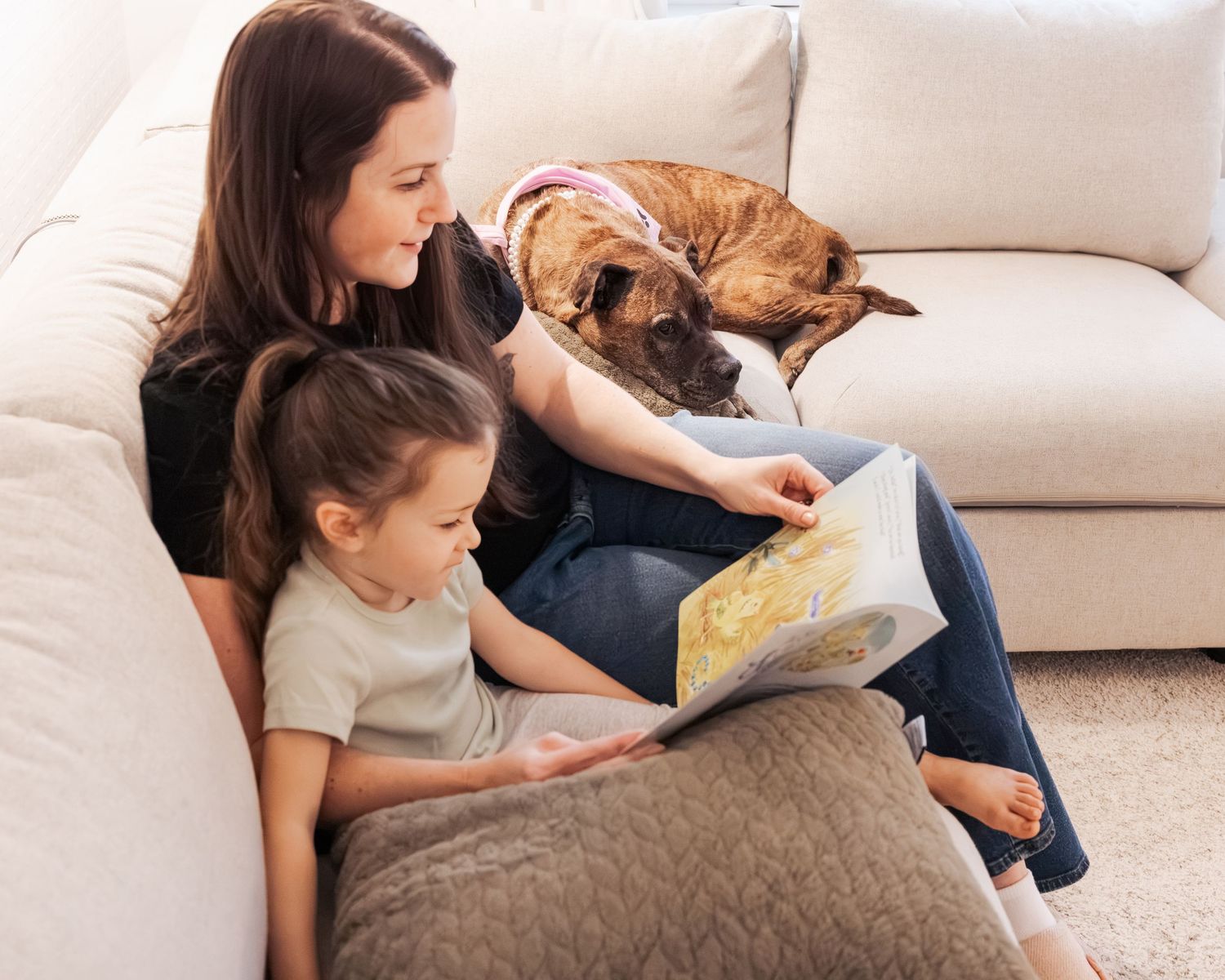 A mother and young daughter read a book together on the floor while a boxer dog watches from the couch.