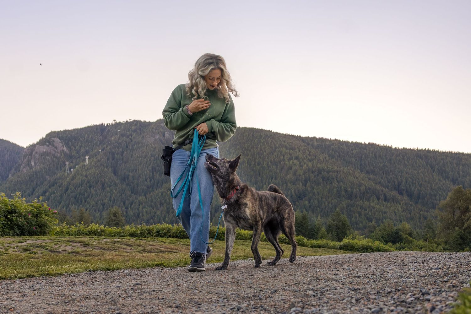 Woman walking with a dark German Shepherd dog on a gravel path with green mountains in the background at dusk.