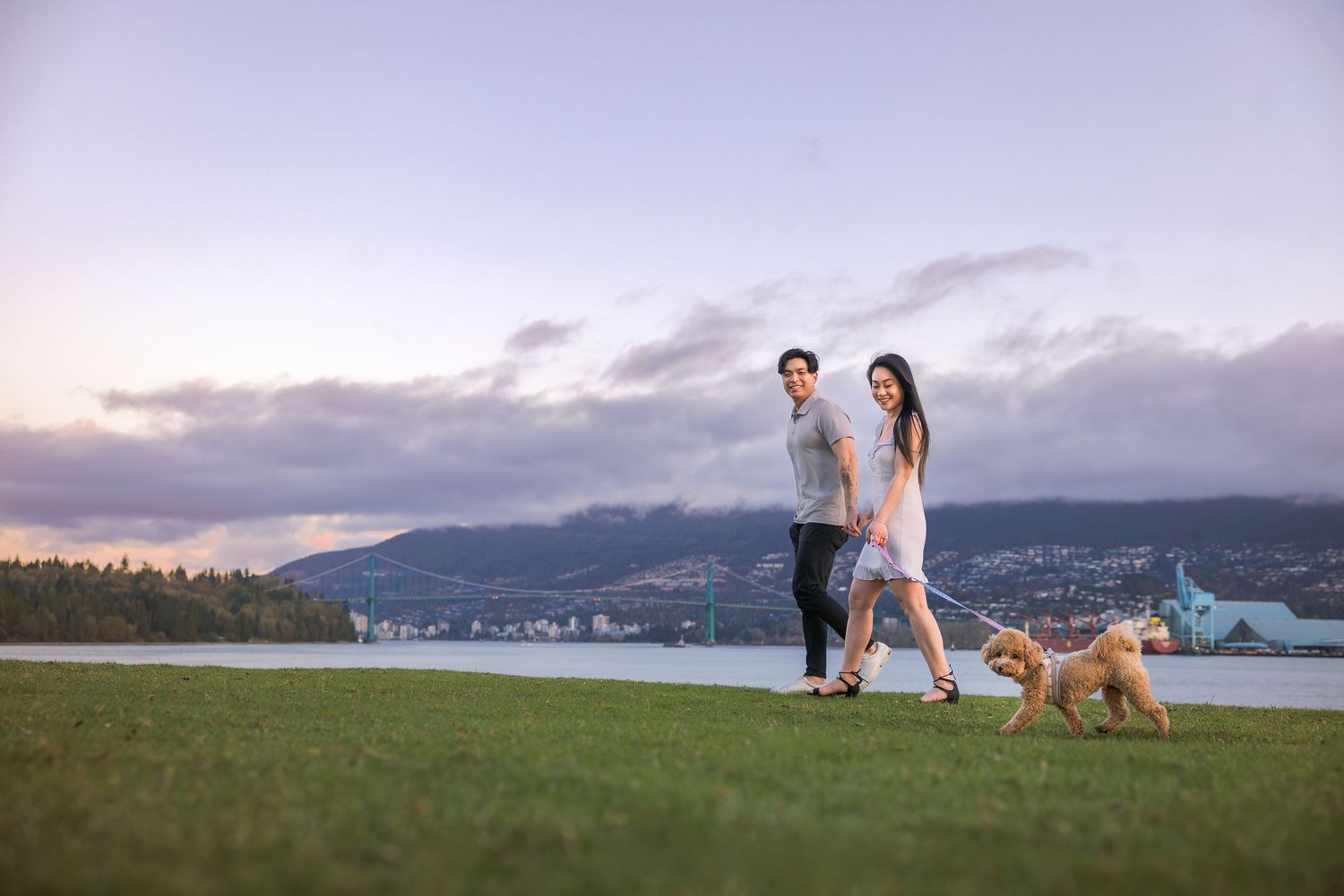 Couple walking a golden dog on green grass by waterfront with mountain backdrop at dusk.