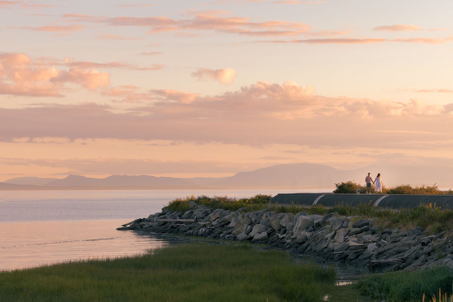 Two people walk along a rocky coastal path at sunset with calm waters and mountains in the background.
