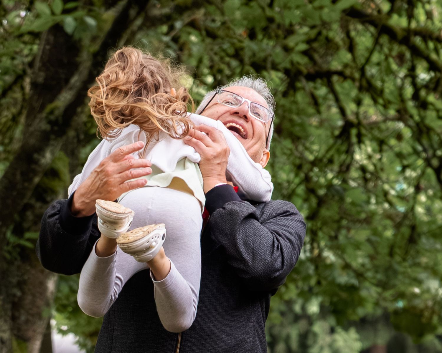 Elderly man joyfully lifts and hugs a young curly-haired child outdoors among lush green trees.