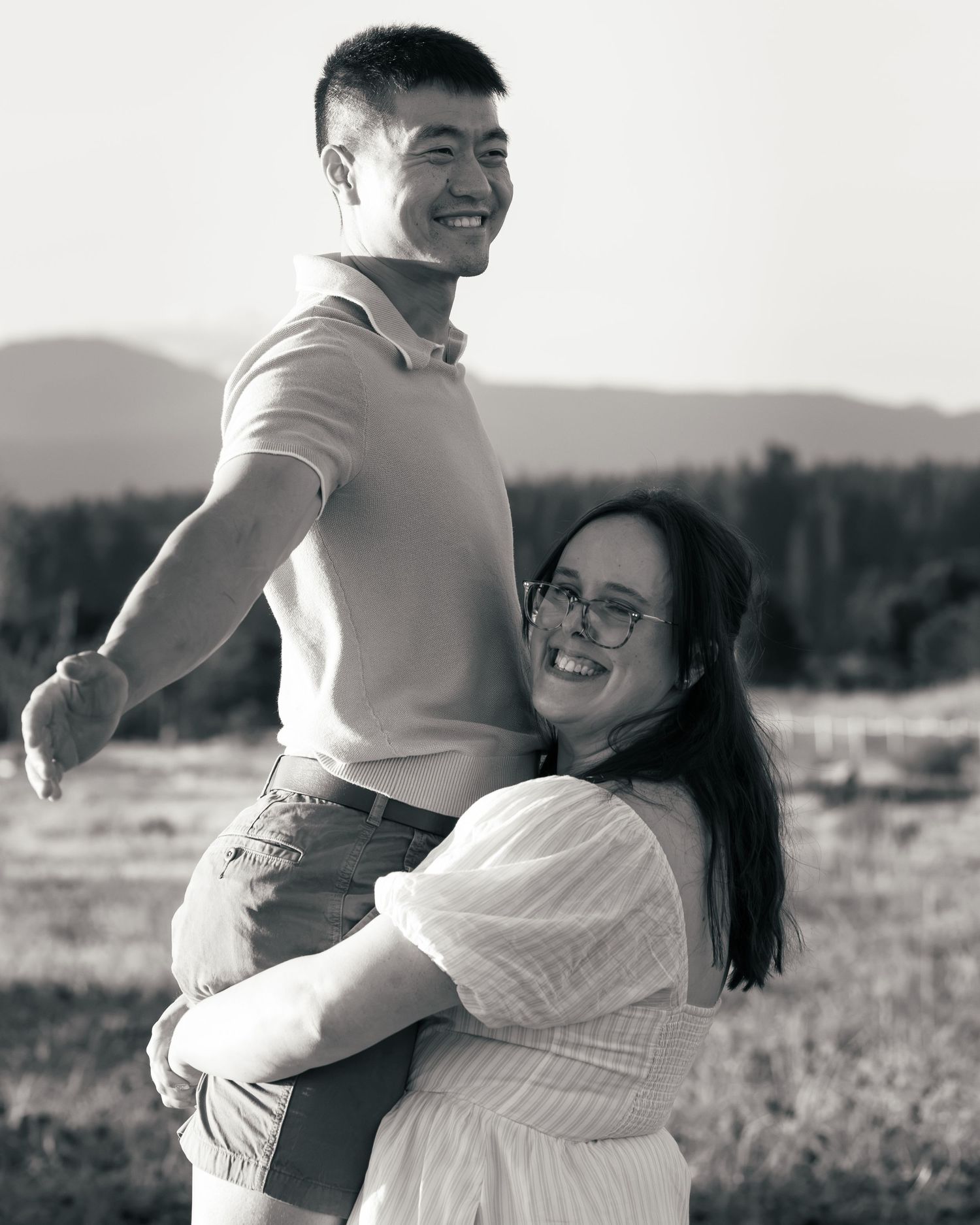 Black and white photo of a smiling couple embracing outdoors with mountains and trees in the background.