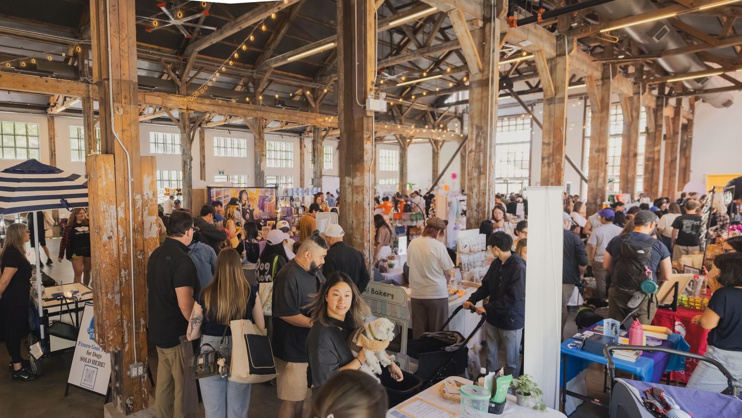 Busy indoor market event with crowds browsing vendor tables in a rustic wooden warehouse venue.