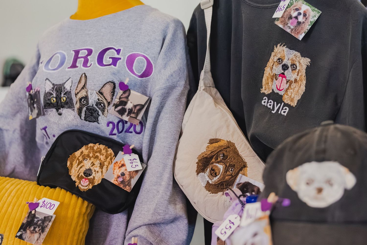 Custom embroidered pet portrait sweatshirts and bags displayed at a vendor booth with dog designs.