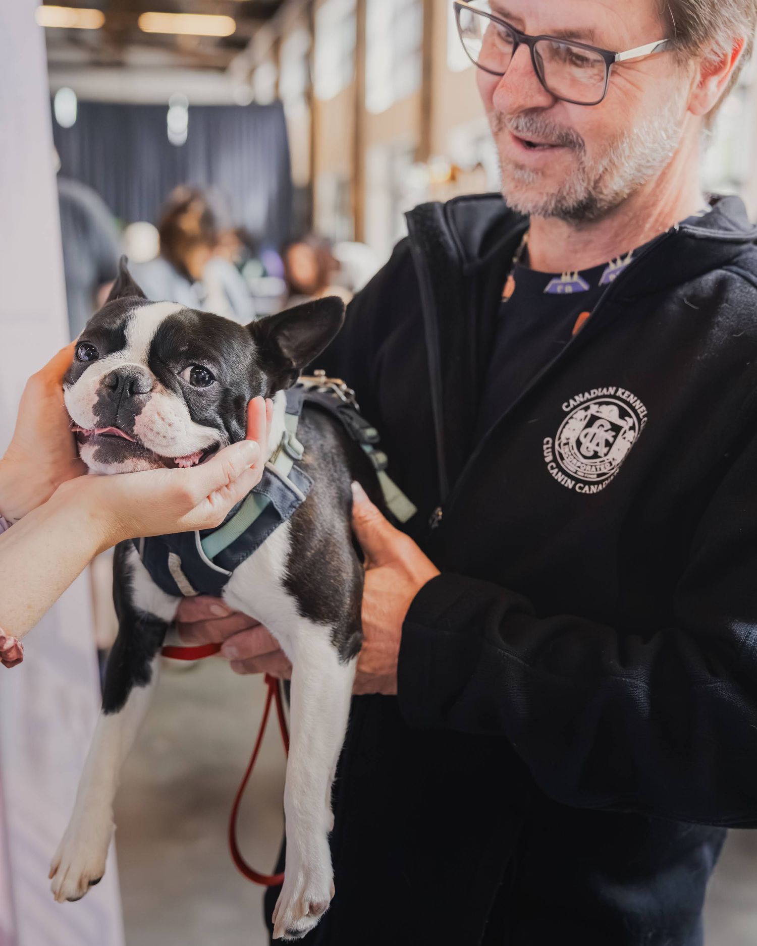 A smiling man in a black jacket holds a happy Boston Terrier dog at an indoor pet event.