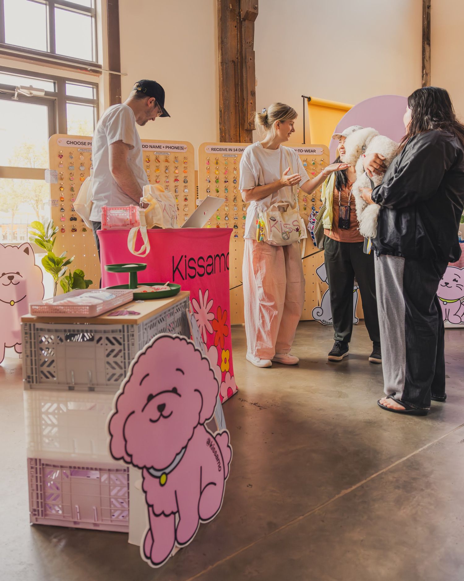 Pink-themed Kissame vendor booth at an indoor market with cute dog cutout display and shoppers browsing merchandise.