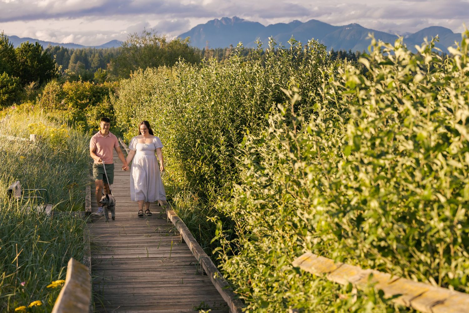 Couple walking hand-in-hand on a wooden boardwalk trail surrounded by lush greenery with mountain views.