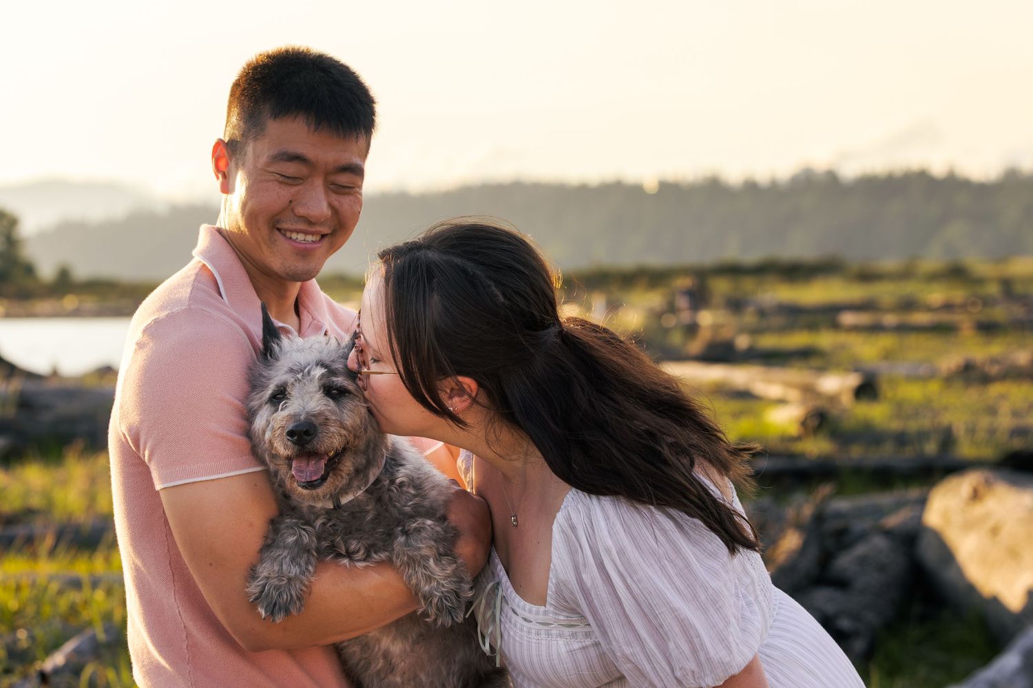 Couple smiling and kissing their fluffy gray dog outdoors at golden hour near a scenic waterfront.