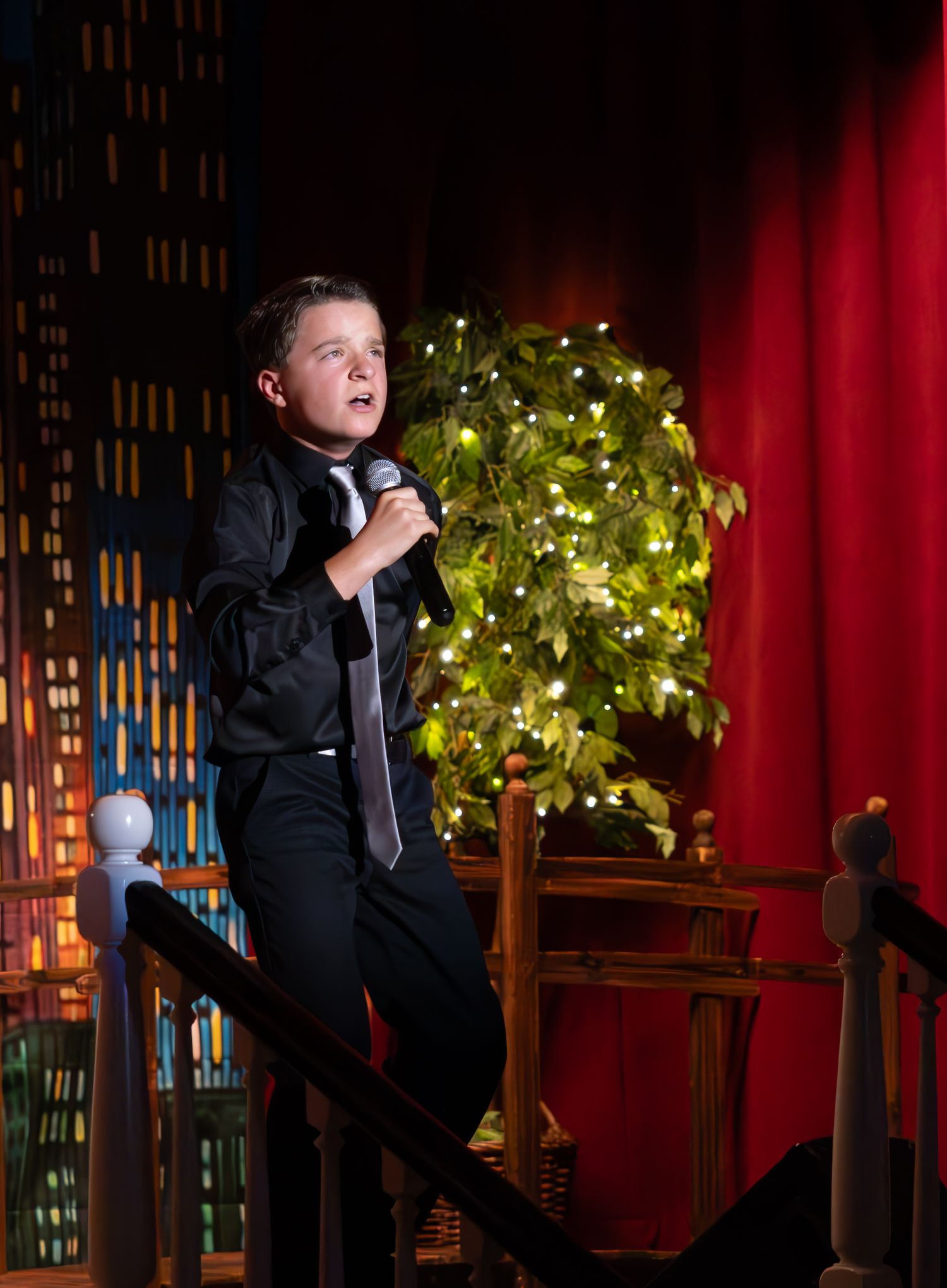 Stage performer in black attire singing against festive backdrop with twinkling lights and holiday decor.