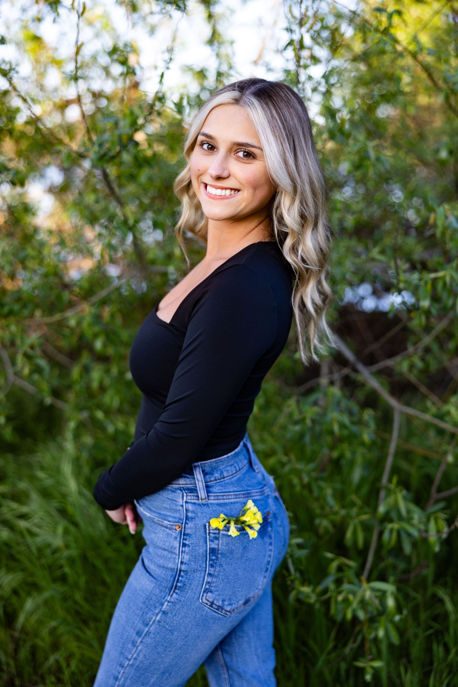 Woman in black top and blue jeans poses outdoors against green foliage background in casual lifestyle photoshoot.
