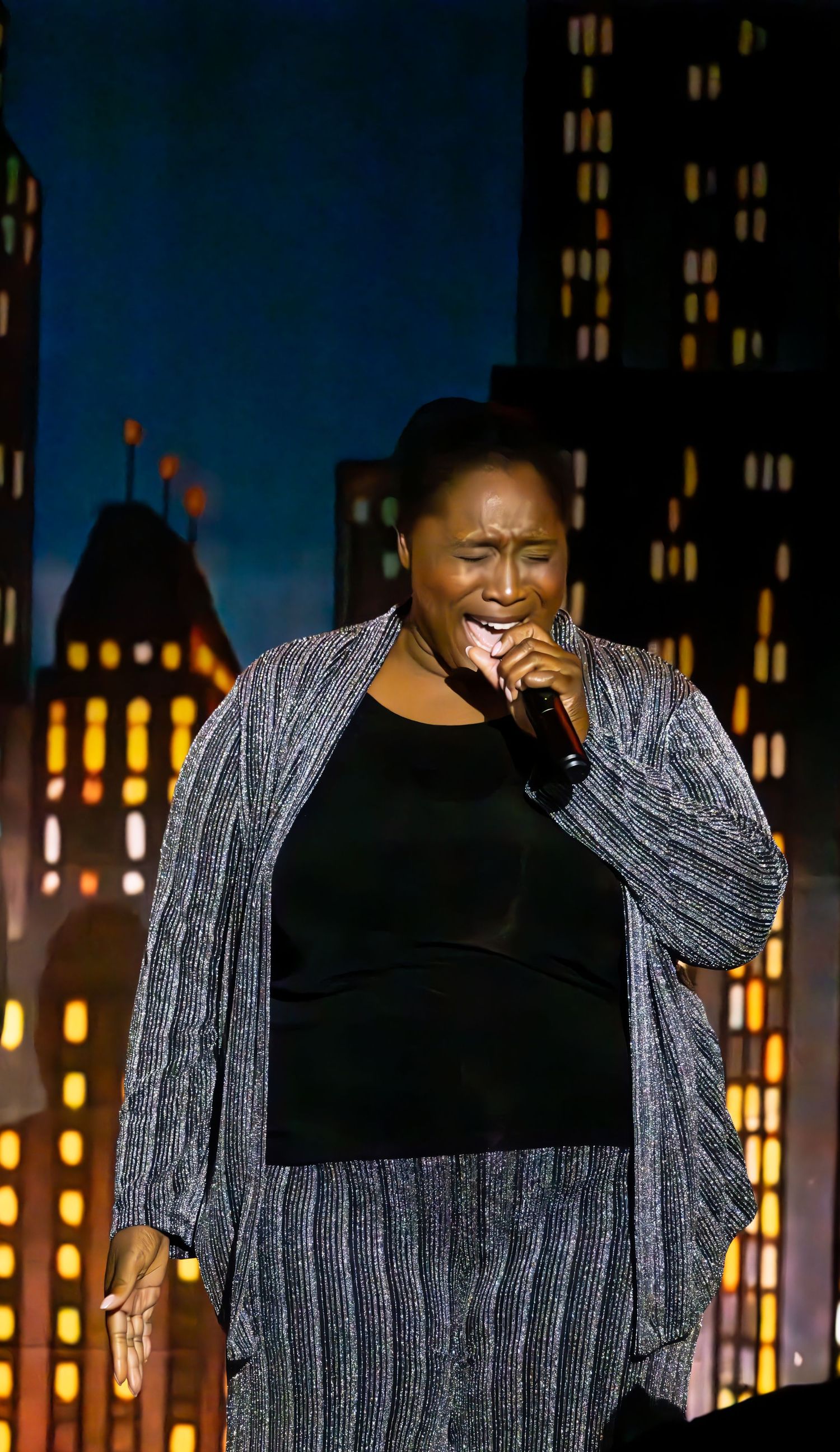 A performer in a glittery gray outfit singing emotively on stage with a cityscape backdrop.