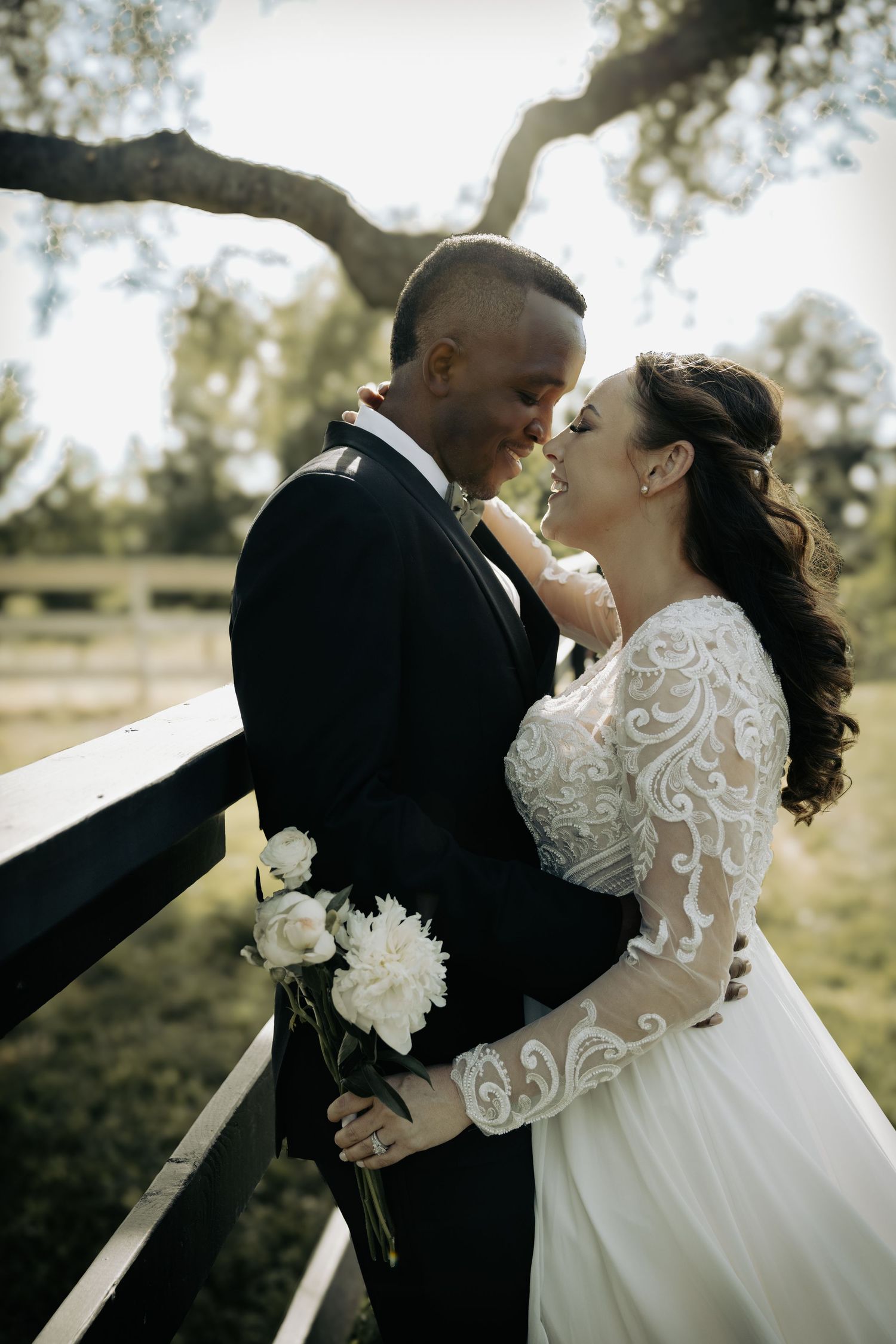 Intimate wedding photograph of newlyweds sharing a tender moment under a tree branch near a rustic wooden fence.