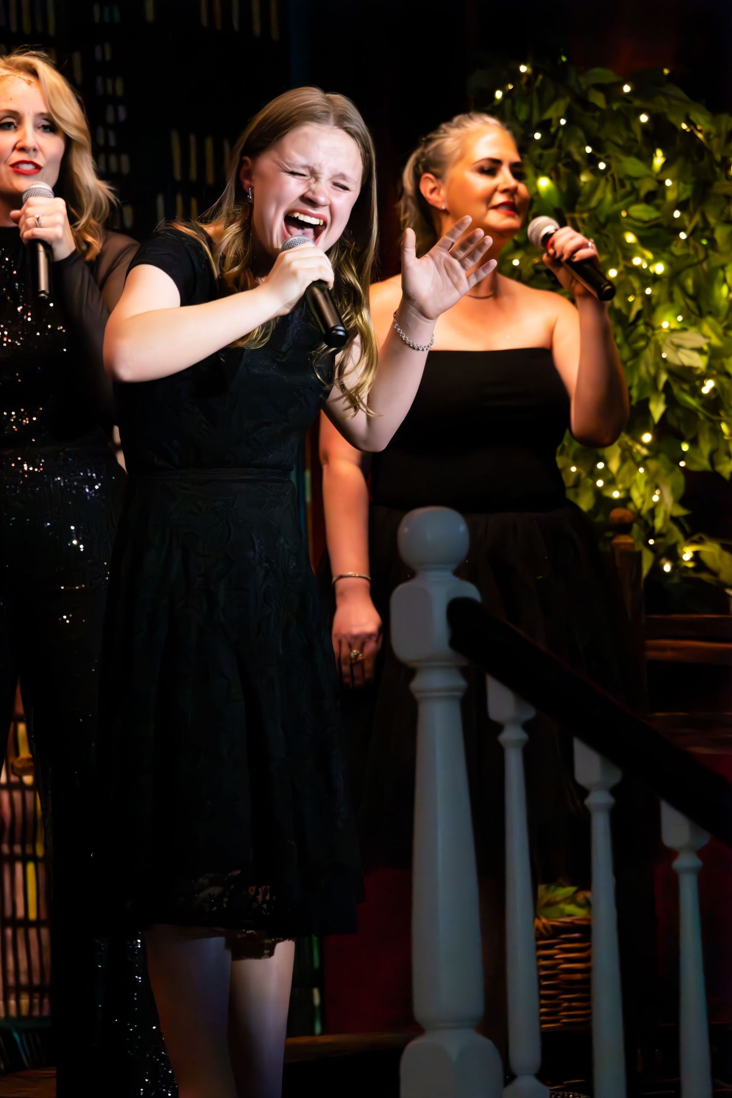A group performs together on a dimly lit stage with white pillars and foliage visible behind them.