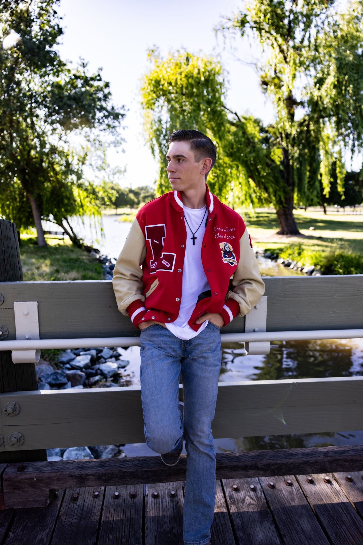 Person in red varsity jacket and jeans sits on white wooden railing by a lake on sunny autumn day.