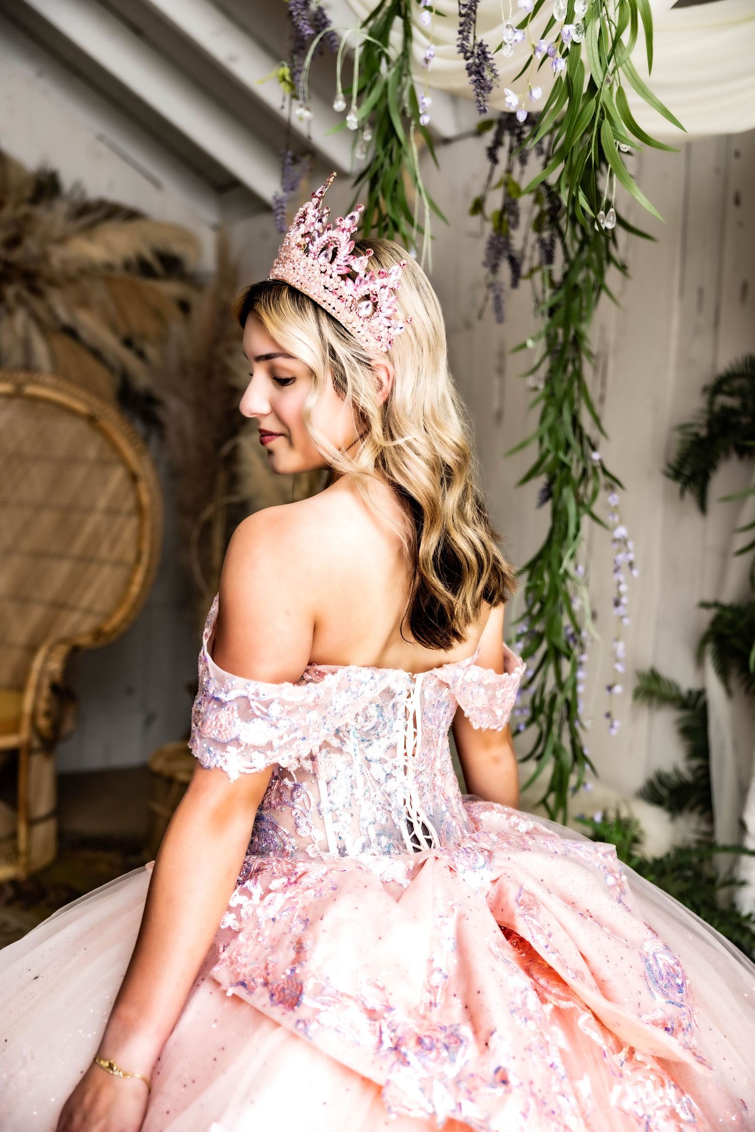 Pink off-shoulder ball gown with floral lace details and tulle skirt in romantic indoor setting with hanging greenery.