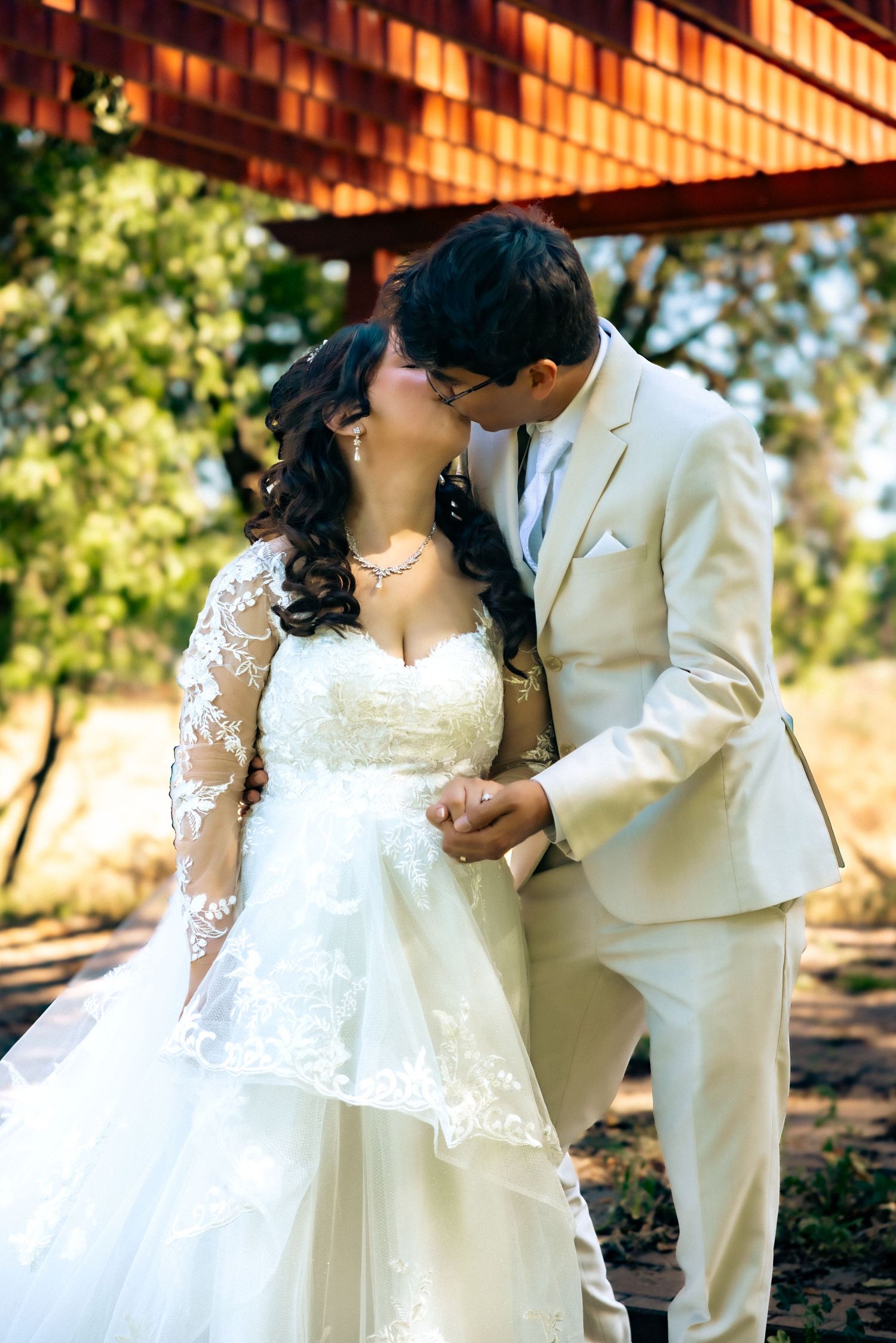 A romantic wedding couple share a tender kiss under a wooden pergola in an outdoor garden setting, surrounded by sunlit greenery.