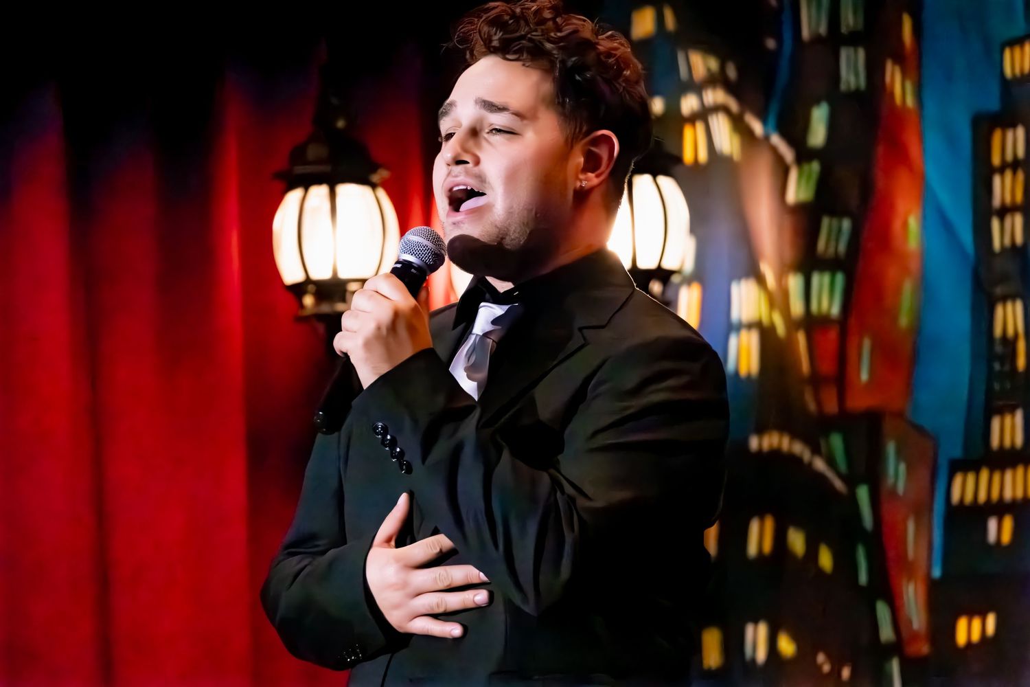 Performer dressed in black stands on stage against city skyline backdrop, gesturing emotionally during show.