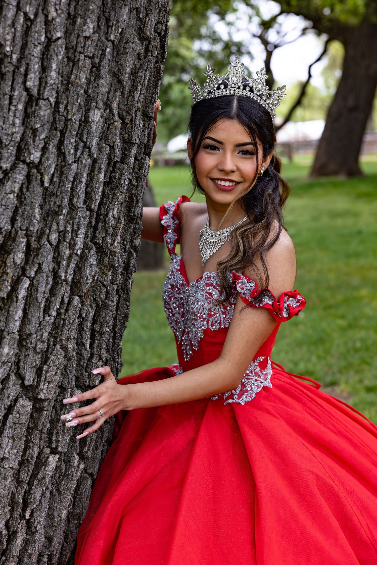 A young woman in a sparkling red quinceañera dress and tiara poses next to a tree trunk in a park setting.