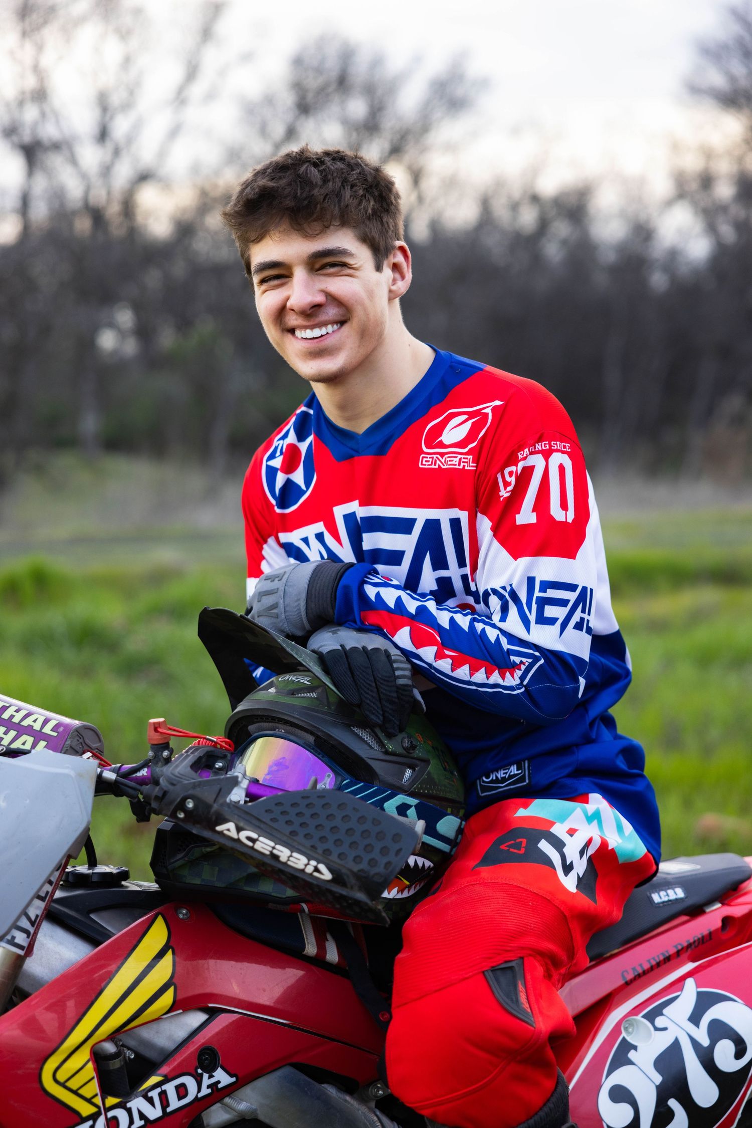 Senior, Motocross rider in red and blue O'Neal jersey poses with dirt bike in grassy field at sunset in Chico for Senior portraits.