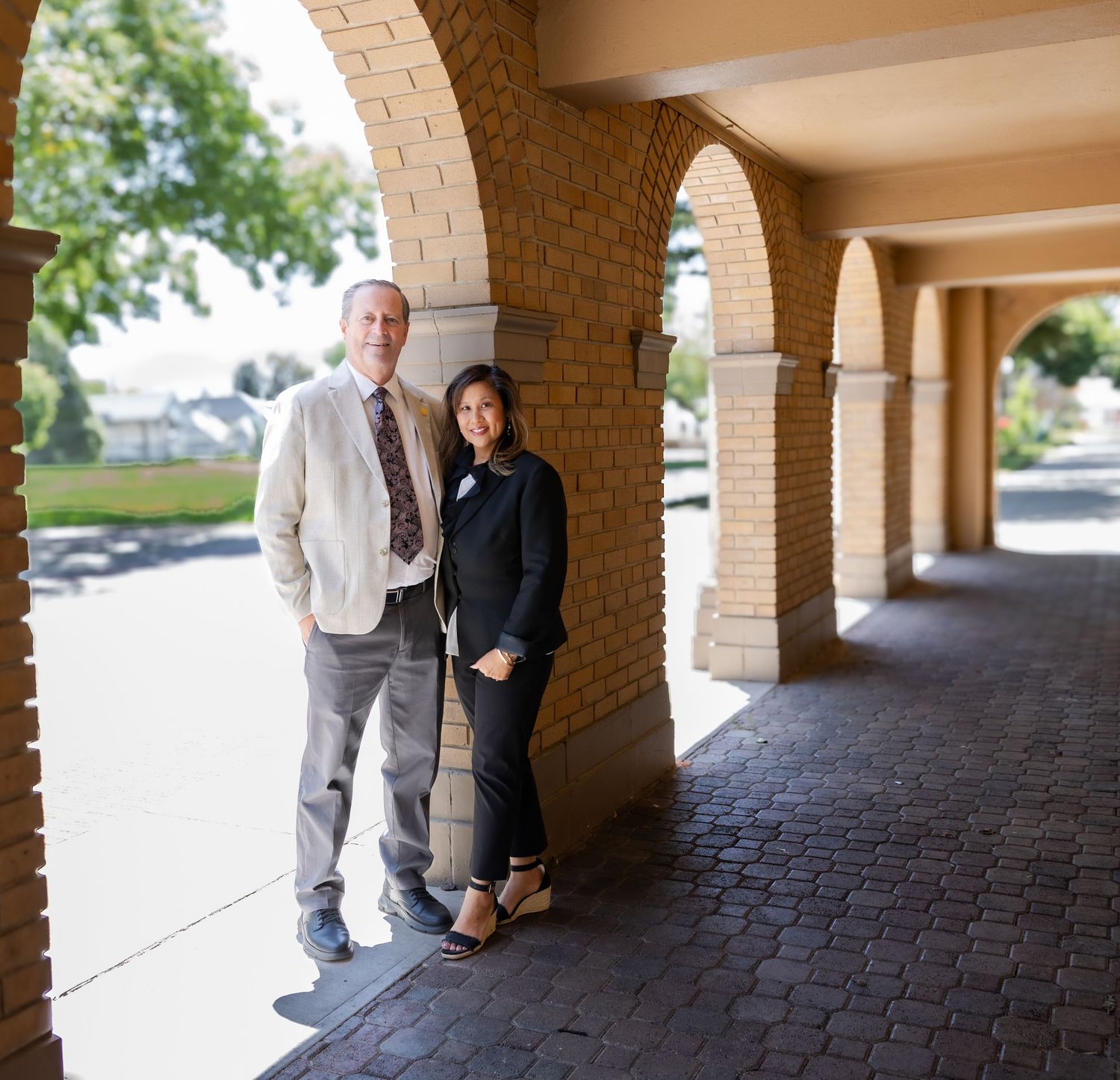 Two people stand in conversation under a covered brick archway walkway on a sunny day.